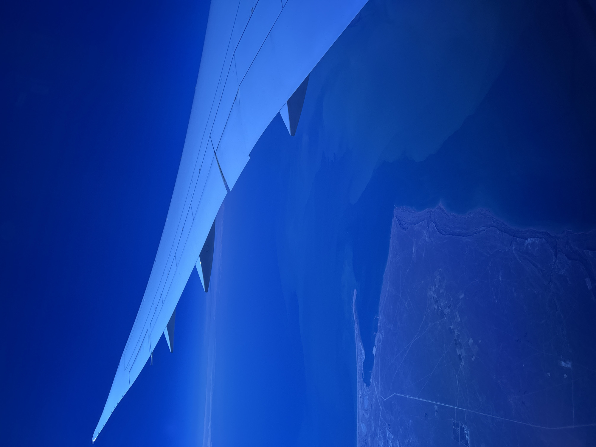 An aerial view shows a portion of an airplane wing extending into the frame from the top left, angled towards the bottom right. The wing is a light blue color and appears to be made of metal with visible panel lines. Below the wing, the vast expanse of a deep blue ocean fills most of the frame. On the left side, a coastline is visible, with a thin strip of land meeting the water. Further inland, the land appears arid and is crisscrossed with faint lines that could indicate roads or irrigation channels. Scattered across this land are several clusters of small, rectangular shapes, possibly agricultural fields or buildings. The overall impression is one of an overcast day from a high altitude.