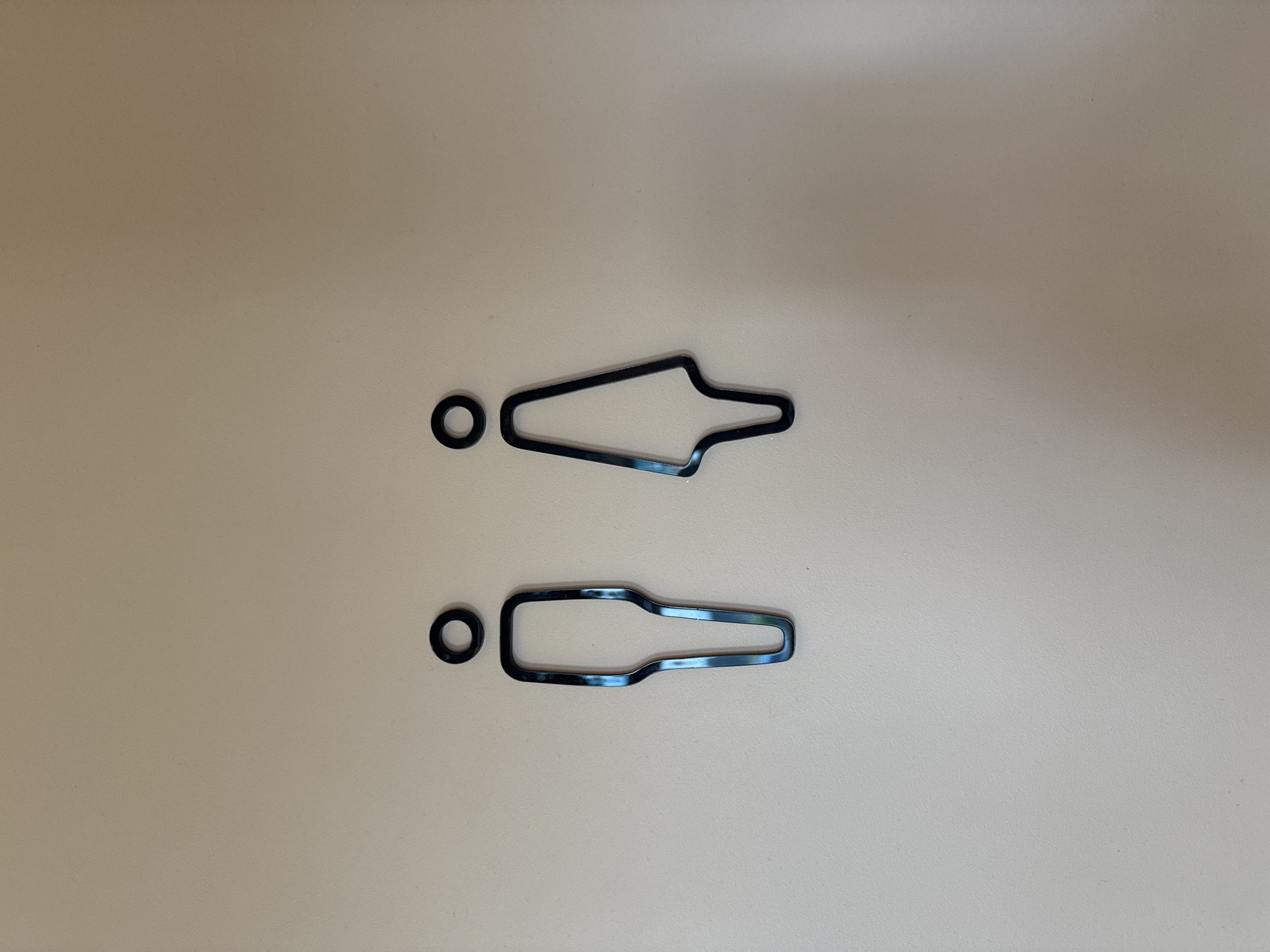 This media file contains a top-down view of two black rubber gaskets on a light beige surface. The gaskets are on the left side of the image. The upper gasket is shaped like a stylized arrow or a tear drop with a pointed end and a wider, rounded end. To the left of this gasket is a small, circular O-ring. Below the upper gasket and O-ring, there is another, larger gasket with a similar shape to the first, but more elongated and with a less pronounced point at one end. To the left of this second gasket is another small, circular O-ring. The lighting is even, suggesting an indoor setting, and there are no shadows that would indicate a specific time of day. There is no visible text or other distinguishing features in the image. The scene appears to be a simple product photograph of the gaskets, possibly for sale or identification. The location context of Патрушева, Russia, does not provide any visual cues within the image itself.