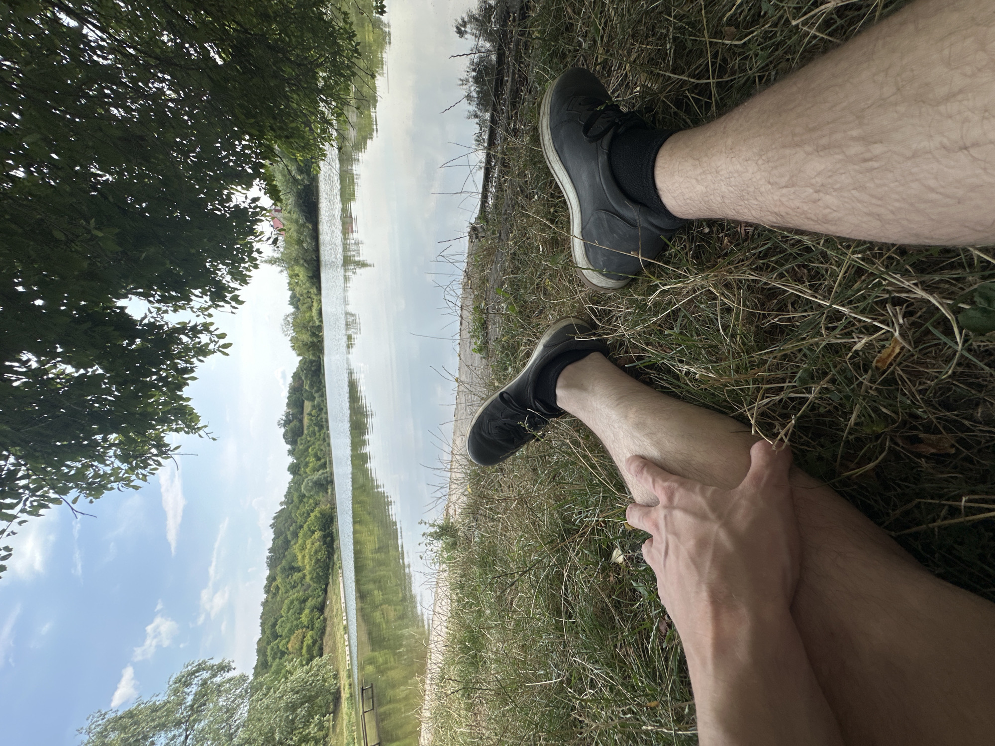 The image shows a low-angle, first-person perspective of a person resting by a lake in Makiv, Ukraine. The person's legs, clad in black shoes and socks, are positioned in the foreground, with one hand resting on the shin of the left leg. The ground is covered in dry grass and some sparse greenery.
Behind the person, a tranquil lake stretches out, reflecting the sky and the surrounding lush green hills covered in trees. The sky is partly cloudy with patches of blue. The atmosphere suggests a peaceful, natural setting, likely a park or recreational area near a body of water. The presence of a wooden structure, possibly a pier or seating area, is visible on the left bank of the lake. A few buildings with red roofs are faintly visible amongst the trees on the far side of the lake. The overall impression is one of relaxation and immersion in nature.