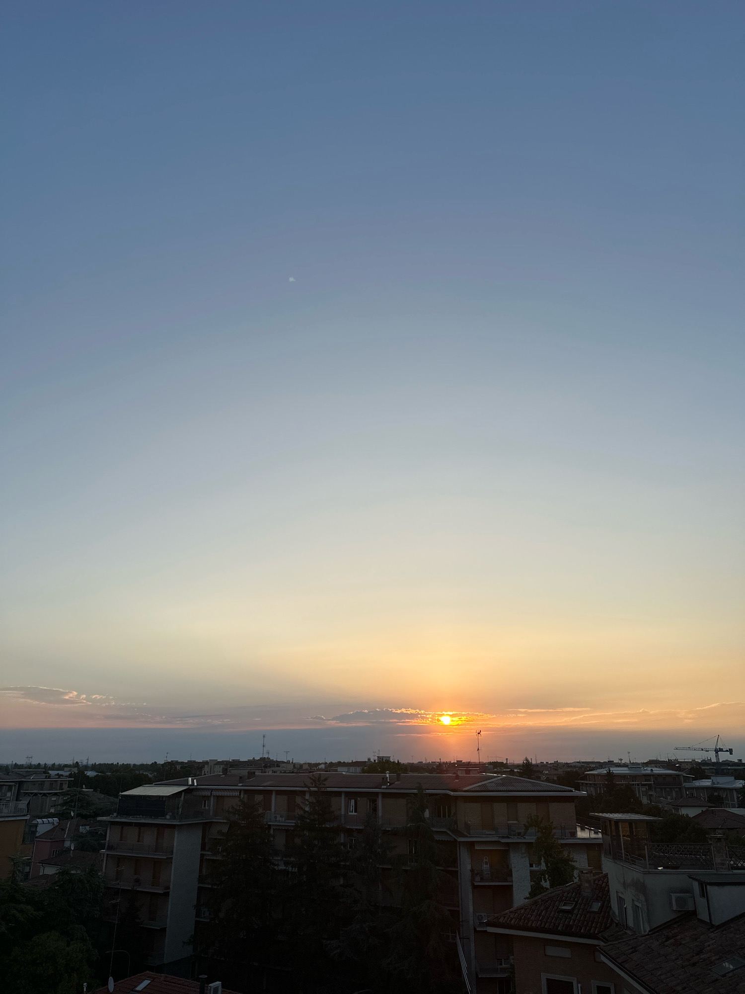A wide shot captures a serene sunset over the cityscape of Parma, Italy. The sky is a gradient of soft blues and pale yellows, with a faint crescent moon visible in the upper left quadrant. The sun, a bright orange orb, is partially obscured by wispy clouds near the horizon, casting long rays of light that illuminate the sky. Below the sky, the rooftops of Parma extend across the frame, featuring a mix of terracotta tiles and flat surfaces. Buildings of varying heights are visible, some appearing residential with balconies and windows, while others suggest commercial or industrial structures. Trees are interspersed throughout the urban landscape, their dark foliage providing a stark contrast to the vibrant sky. The overall impression is one of tranquil urban decay under a beautiful twilight sky.