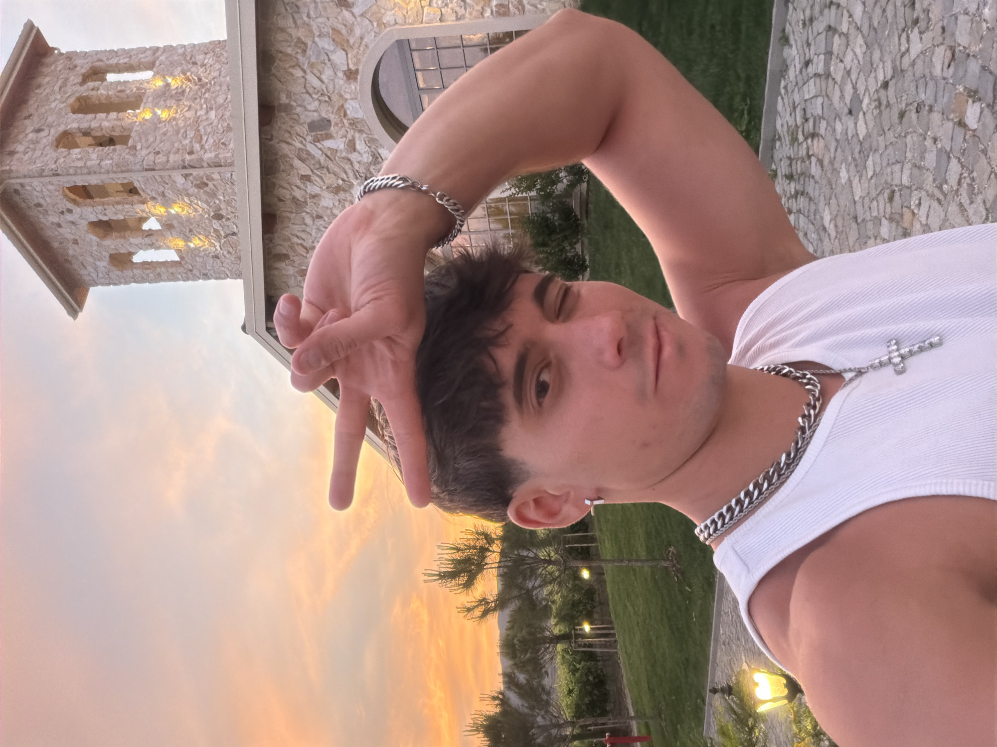 A selfie of a young man is taken outdoors at dusk in front of a stone building with a tower, likely located in Kocherinovo, Bulgaria. The man is wearing a white tank top, a thick silver chain with a cross pendant, and a silver bracelet on his right arm. He has dark hair and is winking with his left eye while holding his right hand up to his forehead with his index and middle fingers forming a peace sign. The sky behind him is a gradient of orange and pink, indicative of a sunset. The building features stone construction and arched windows, with lights illuminating the tower. The foreground shows a cobblestone path and green grass.