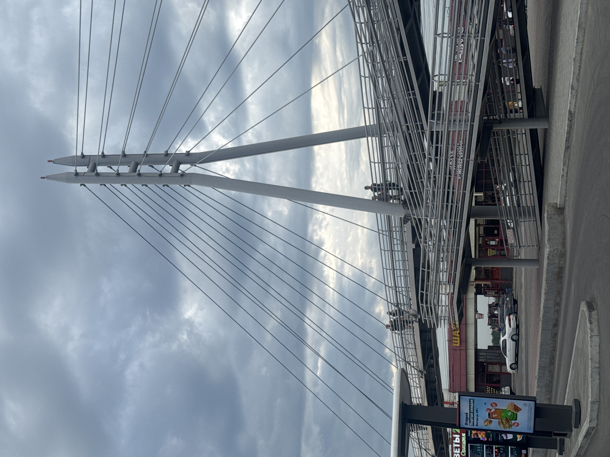This is a medium-angle, eye-level shot of a pedestrian overpass in Irkutsk, Russia. The overpass is a modern, cable-stayed structure with two tall, white support towers. Numerous grey cables connect the towers to the walkway. The walkway itself is constructed of grey metal, with ramps leading up and down on either side. Several people are visible on the walkway, walking in both directions. Below the overpass, there is a street with two cars parked and another moving. Behind the street, there are several storefronts with red and white awnings. The sky is cloudy and overcast, suggesting it may be a cooler day. The overall impression is one of an urban, functional space.