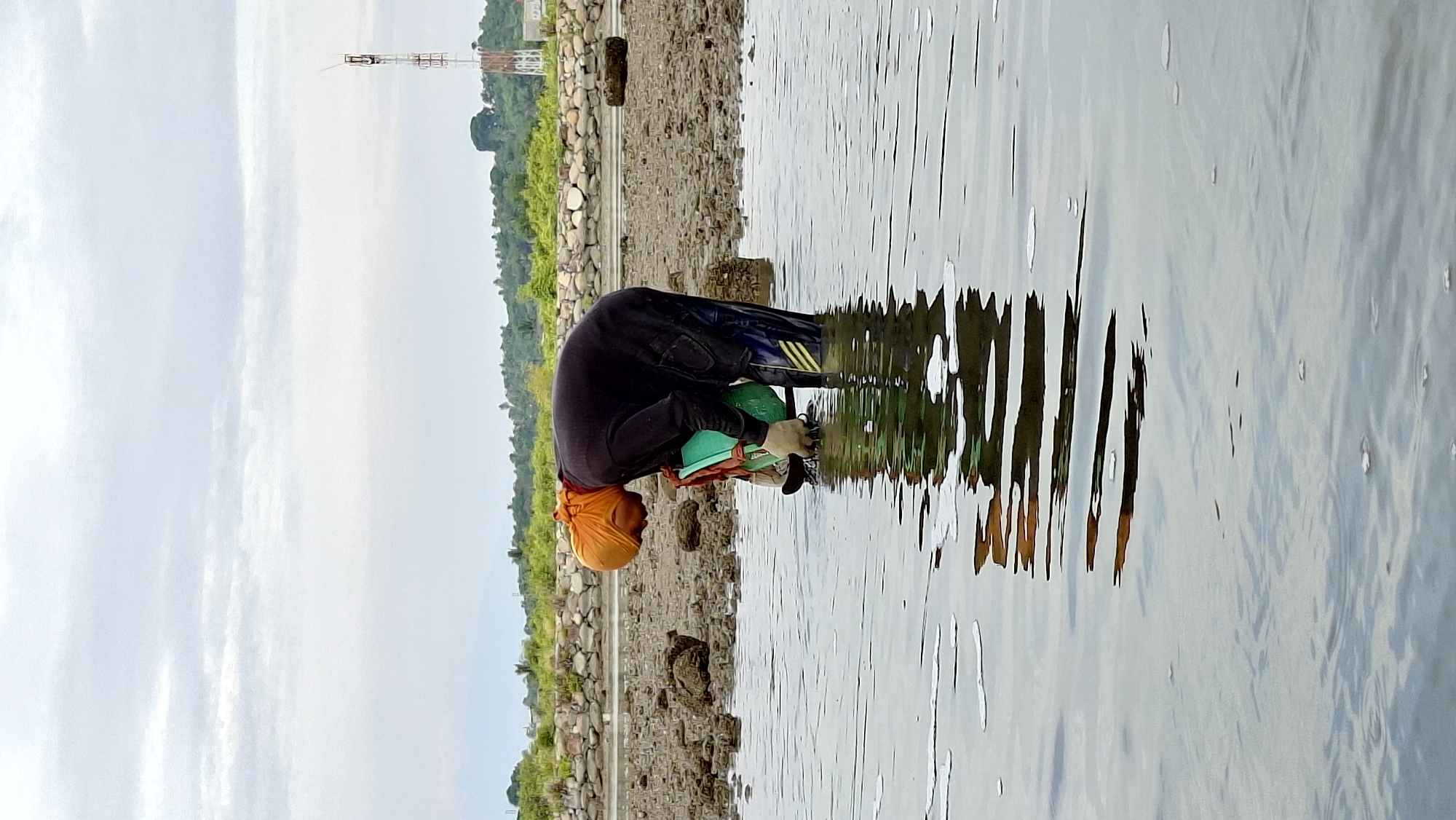 An individual, wearing an orange head covering, dark long-sleeved shirt, and dark pants with yellow stripes, is bent over collecting items from shallow water. They are wearing gloves and holding a green bucket. The scene takes place on a muddy shore with scattered rocks and vegetation in the background, under a cloudy sky. A telecommunications tower is visible in the distance, suggesting an urban or semi-urban coastal environment. The location is likely Lhokseumawe, Indonesia, a city known for its coastline and industrial presence.