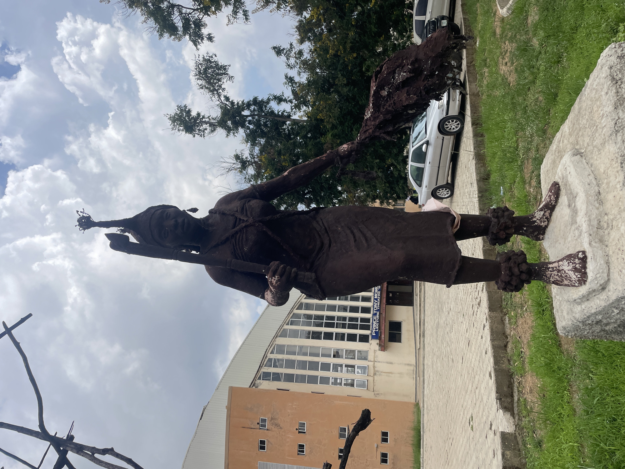 The media file presents an outdoor scene featuring a dark brown statue of a person, likely male, standing prominently on a light-colored base. The statue depicts a figure wearing a headdress, a necklace, a skirt-like garment, and anklets. It holds a staff or spear in its left hand and an object resembling a fly-whisk or ceremonial item in its raised right hand. The statue shows some signs of weathering or paint erosion, with lighter specks on its surface, particularly on the base and feet.
In the mid-ground, a large, light-colored building with a distinctive arched roof and numerous rectangular windows is visible. A prominent sign on this building reads "PRINCE TONY MOMOH LECTURE THEATRE 'B'". Behind and to the left of this building stands another smaller, light brown structure with multiple square windows.
To the right of the statue, two white sedans are parked on a paved area, which appears to be a road or parking space. Green grass covers the ground in the foreground and borders the paved areas. Scattered trees are visible in the background and upper frame. The sky is partly cloudy with white and grey clouds, indicating daytime. The presence of the lecture theatre sign strongly suggests an academic or institutional setting, likely a university campus in Jos, Nigeria. No direct human activity is taking place; the scene is static.