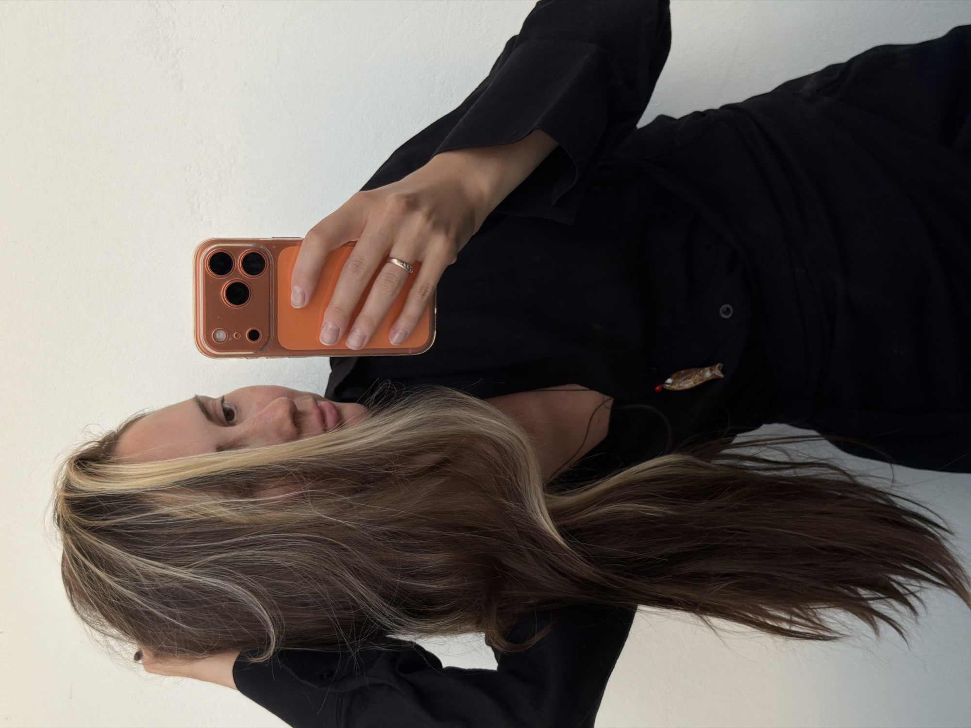 A sideways, medium shot captures a person, likely a woman, in an indoor setting against a plain, light-colored wall. The individual has long, wavy brown hair with prominent blonde highlights, and fair skin with visible freckles on her face. She is wearing a black long-sleeved top or dress.
The person is holding a smartphone in her left hand, which is adorned with a ring on the fourth finger. The phone has an orange case with a clear bumper, clearly showing a camera module with three main lenses and a smaller sensor. Her gaze is directed towards the phone screen, suggesting she is interacting with the device, possibly looking at content or preparing to take a photo/video.
Notable details include a small, decorative brooch resembling a stylized fish or insect pinned to the black clothing near her left shoulder. The overall impression is one of casual interaction with a personal device. The scene takes place in Khimki, Russia. There is no visible text in the image.