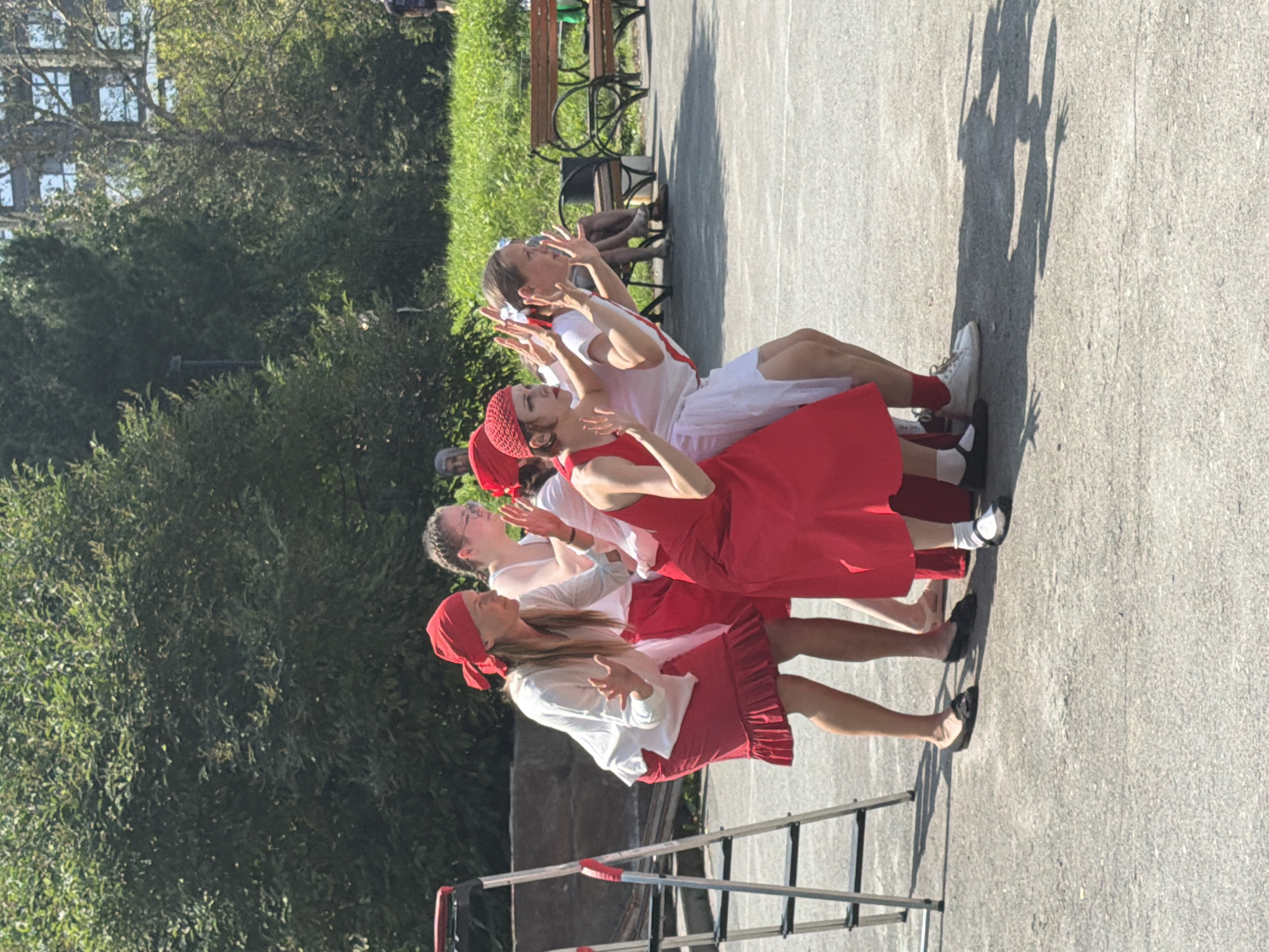 A group of five women, dressed in predominantly red and white outfits, are performing a choreographed dance outdoors in Yekaterinburg, Russia. The women are facing away from the camera, with their hands raised in front of them, as if in unison. Three of the women are wearing red headscarves. The woman in the front left is wearing a white long-sleeved shirt and a red skirt. The woman to her right is wearing a red outfit. The woman in the center front is wearing a red dress and a red headband. The woman to her right is wearing a white top with red accents and a white skirt. The woman on the far right is wearing a white top with red accents and a white skirt.
The performance is taking place on an asphalt surface, likely a park or public square. To the left of the group, a silver ladder with red accents is leaning against a low wall. In the background, there are lush green trees and a glimpse of a building. The lighting suggests it is a sunny day, casting distinct shadows on the ground. Two park benches are visible in the background to the right, with some individuals sitting on them. The overall impression is of a community event or performance in an urban green space.
