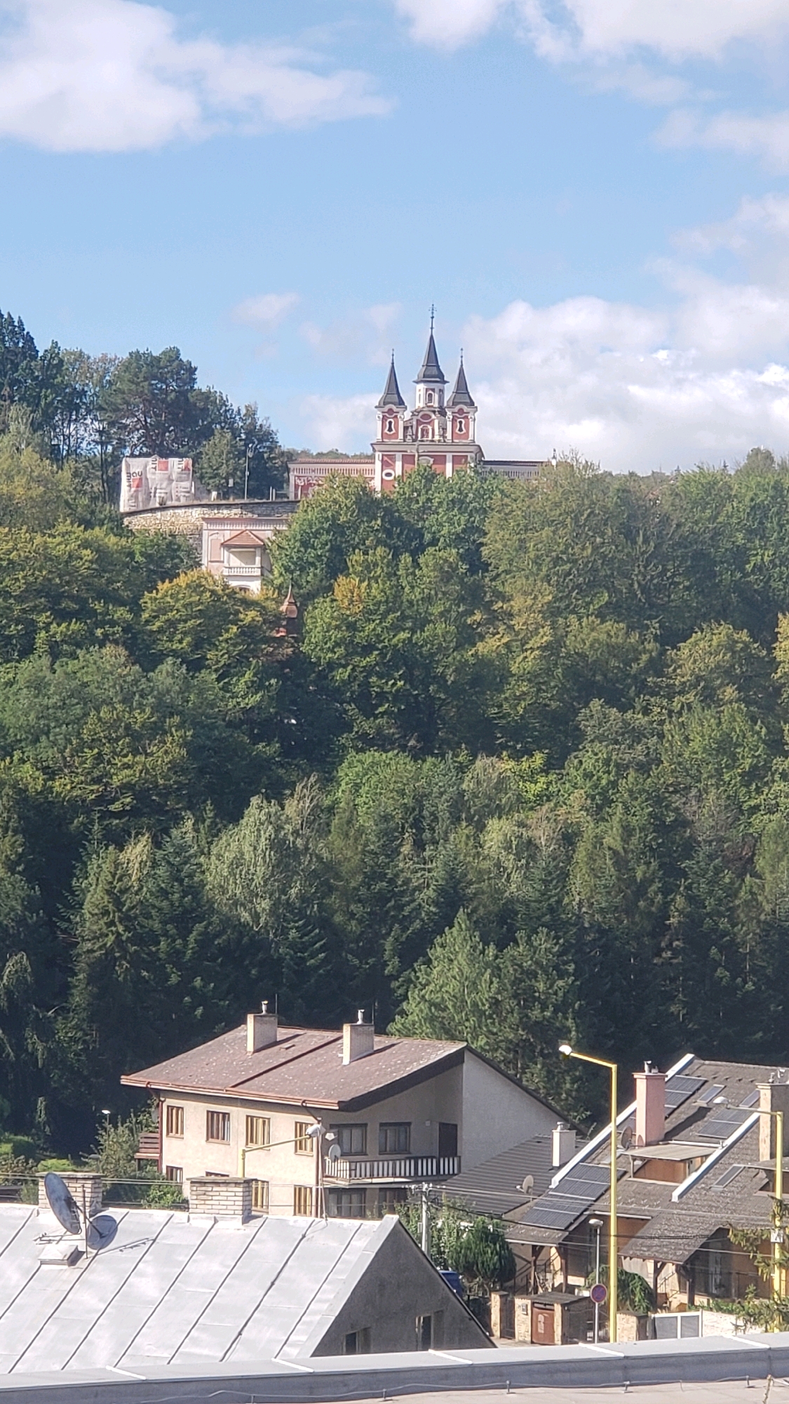 The image depicts a view of the Holy Trinity Church and Monastery in Prešov, Slovakia. The church, with its distinctive red and white facade and multiple spires, is situated atop a densely forested hill. Below the church, closer to the foreground, are several residential buildings, including a light-colored house with a dark roof, a house with solar panels, and other structures. The roofs of these houses are visible, some featuring chimneys and satellite dishes. The scene is set against a backdrop of lush green trees and a partly cloudy blue sky. The overall impression is one of a town nestled within a natural landscape, with a prominent religious structure overlooking the residential area.