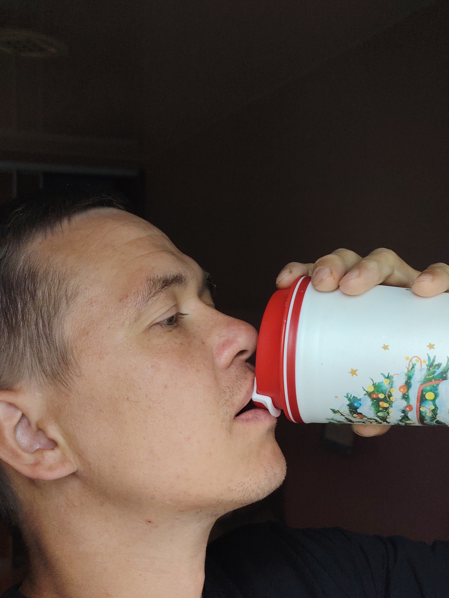 A medium shot shows a man drinking from a white, cylindrical container with red trim and a Christmas tree design. The man's face is turned to the right, and he is in profile. His eyes are closed, and his lips are pressed against the spout of the container. His hand is visible, holding the container. The background is dark and appears to be indoors. There is no information in the image to suggest it is located in Chaikovsky, Russia.