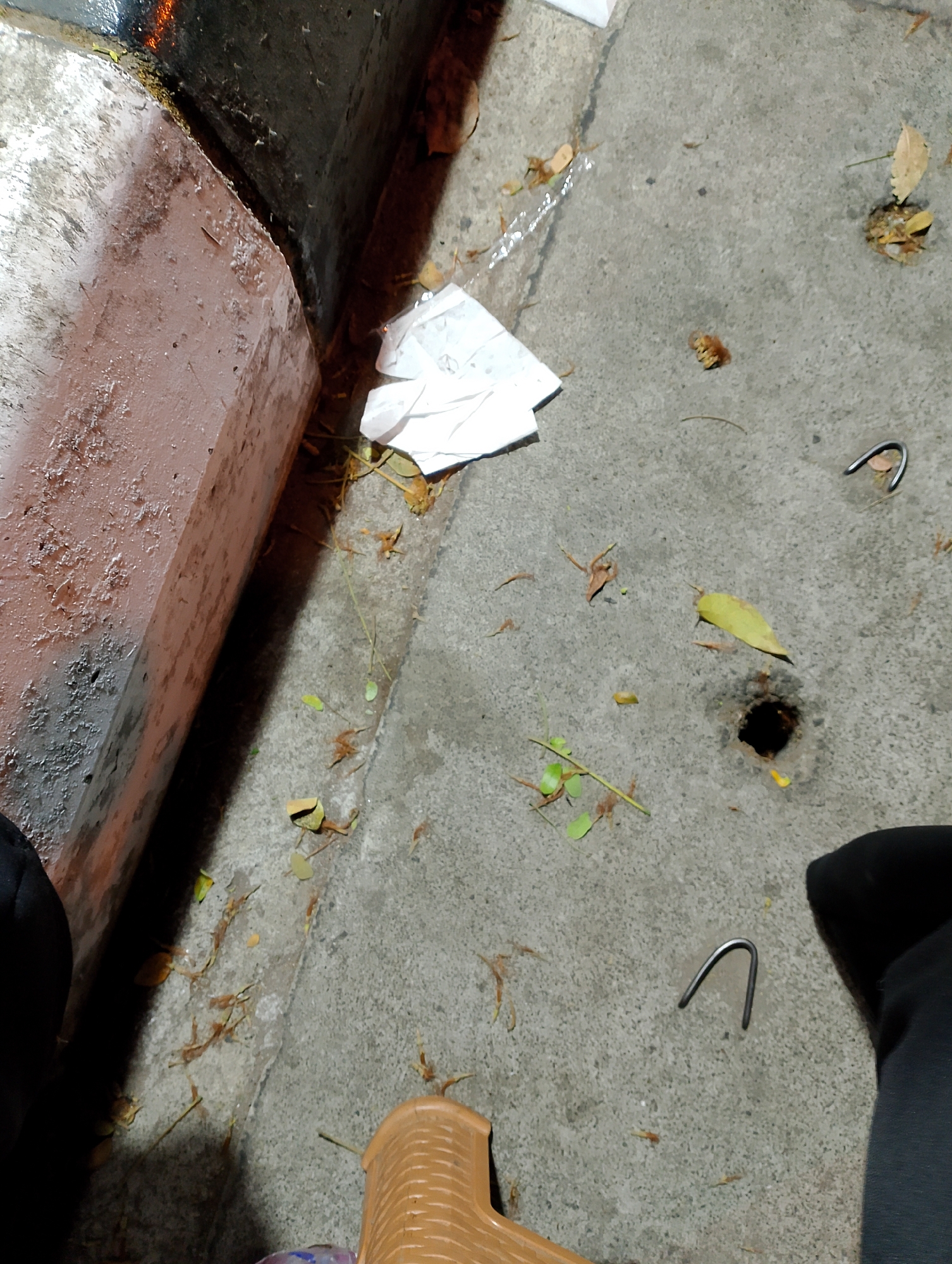 The image depicts a ground-level view of a concrete surface with a reddish-brown, weathered structure to the left. The ground is scattered with dry leaves, twigs, and a crumpled piece of white paper. Two U-shaped metal objects are visible on the concrete. A dark, circular hole is also present in the ground. In the lower right and left corners, parts of dark clothing and a light brown plastic chair are visible, suggesting the presence of at least one person. The overall scene suggests an outdoor environment, possibly a sidewalk or street, with signs of neglect and debris. The location in Magelang, Indonesia, indicates this scene is likely an urban outdoor setting within that city.