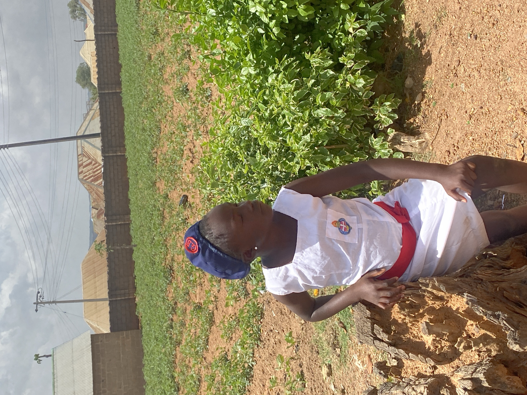 A person wearing a white uniform, red sash, and navy blue cap with the letters "G. B." visible on the cap lies on a weathered tree trunk in Turu, Nigeria. The individual is positioned in a field with sparse vegetation. In the background, there are buildings with corrugated metal roofs and block walls under a cloudy sky. A patch of dense green foliage is located near the tree trunk. A power pole and lines are visible against the sky. The ground is dry and appears to be dirt or sand.