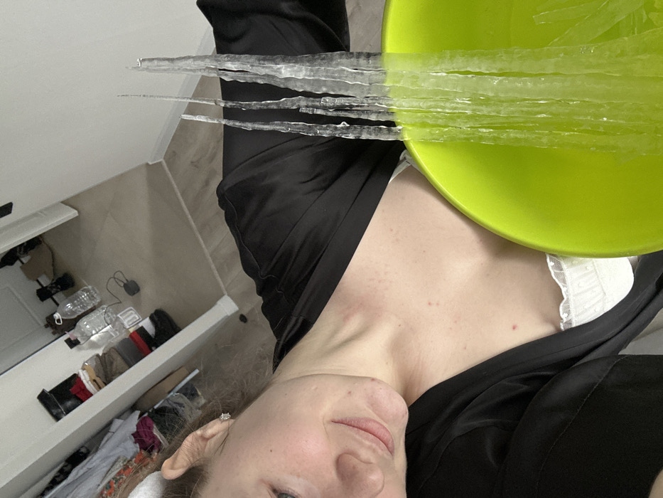A person faces the camera, a green bowl hovering over their chest. Long, translucent strips rest on the bowl's edge, extending like icicles. The person wears a black garment over a white top with lace detailing. The background shows a lightly colored room with a partially open dresser. Various items, including water bottles and clothing, are visible in the drawer.