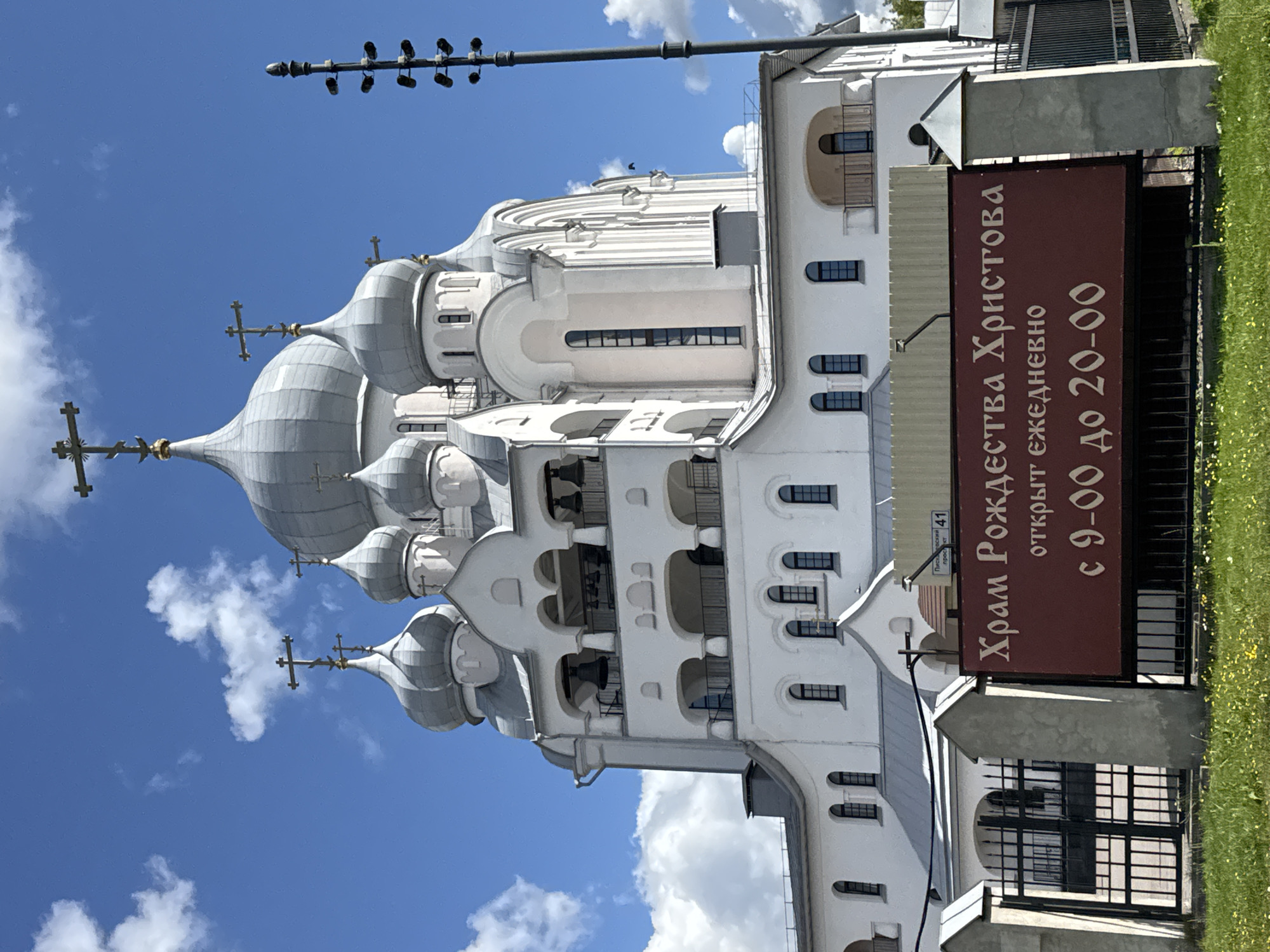 The image shows the Church of the Nativity of Christ, located in Saint Petersburg, Russia. The church is a large, white building with multiple domes topped with crosses. The main dome is silver, and there are several smaller domes also in silver. The building features arched windows and balconies, and a bell tower with bells visible. In front of the church, there is a dark red sign with text in Cyrillic. The sign reads "Храм Рождества Христова" (Church of the Nativity of Christ) and "открыт ежедневно с 9-00 до 20-00" (open daily from 9:00 to 20:00). A black metal fence is visible in front of the sign, and beyond that, a black wrought-iron gate. The foreground shows a grassy area with small yellow flowers and a portion of a grey concrete fence. A tall, multi-light lamp post stands to the right of the church. The sky is bright blue with scattered white clouds. There are no people visible in the image.