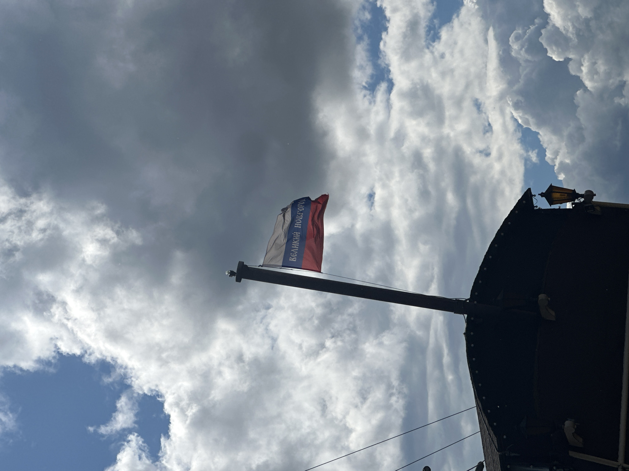 A flag with blue, white, and red horizontal stripes and blue Cyrillic text is affixed to a dark pole. The flag reads "ВЕЛИКИЙ НОВГОРОД", which translates to "Veliky Novgorod". The pole extends upwards from the dark, curved roof of a structure that appears to be a building or a boat. The structure has several small lights along its edge and a vintage-style lantern attached to its side. The scene is set against a cloudy sky with patches of blue visible. The overall impression is an outdoor setting in Veliky Novgorod, Russia.