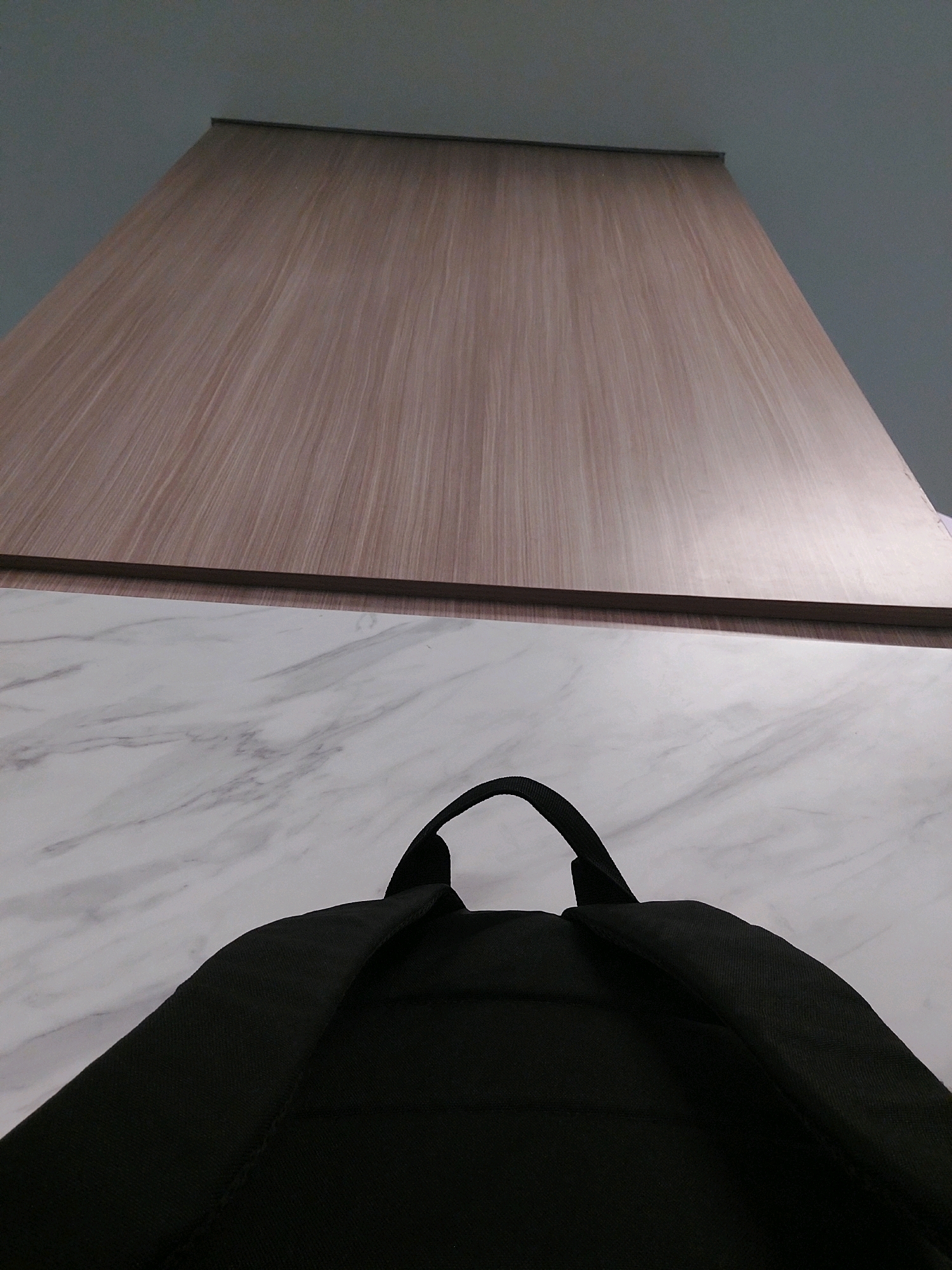 The image shows a low-angle view of a black backpack from behind, placed on a marble-patterned surface. Above the backpack and surface, there is a wooden panel with a vertical grain texture, and above that, a plain gray ceiling. The scene appears to be indoors, possibly in an office or home setting. There are no people or specific landmarks visible that would indicate a location in Bekasi, Indonesia.