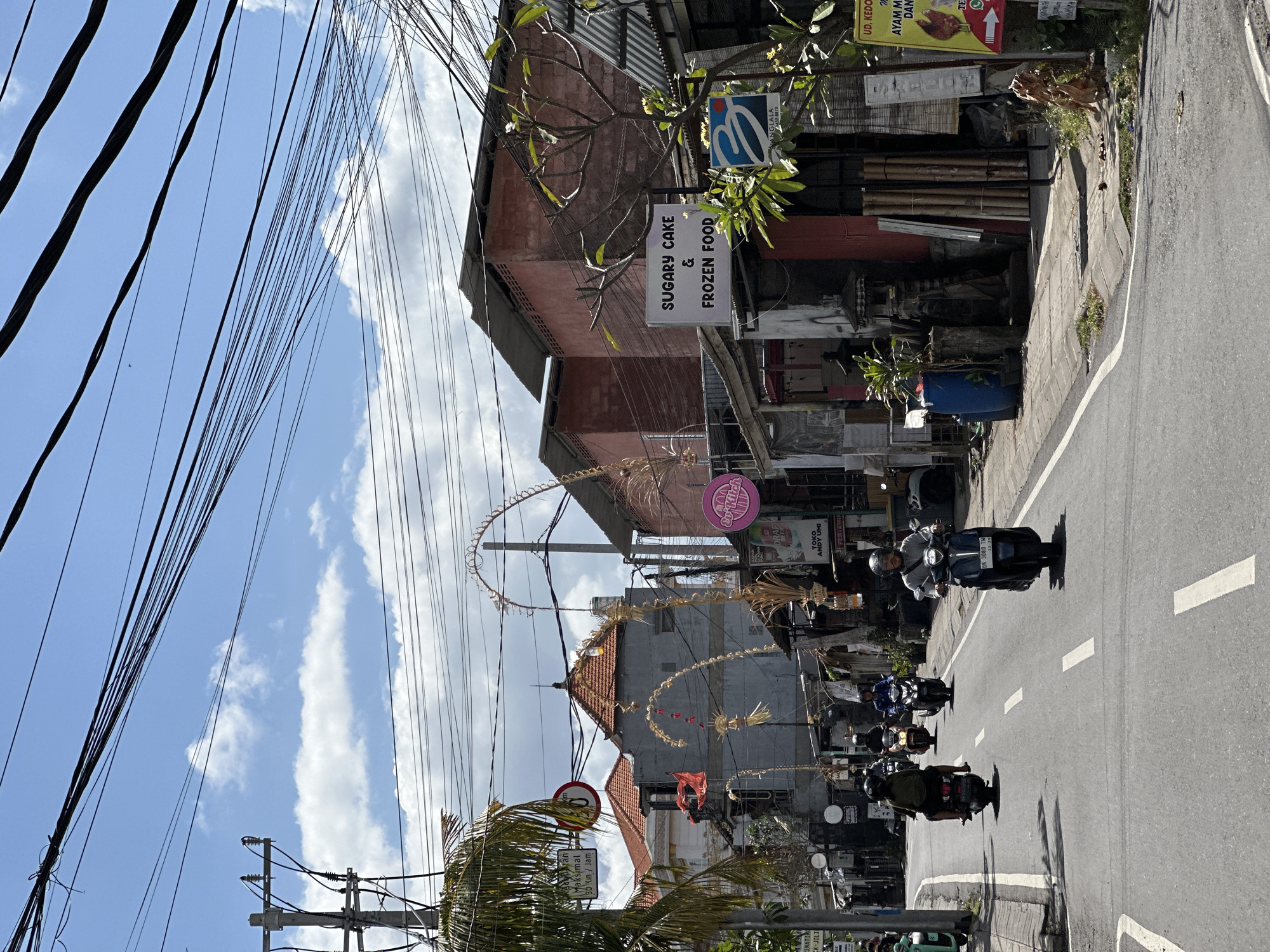 This image depicts a street scene in North Kuta, Indonesia, on a sunny day with a bright blue sky dotted with white clouds. The street is paved with asphalt and marked with white dashed lines. Several motorbikes are visible, with riders on them, moving along the street. Some are in motion, while others appear to be stopped or moving slowly.
On either side of the street are buildings with various storefronts. One sign reads "SUGARY CAKE & FROZEN FOOD". Another sign features a blue logo with the letter "B". There are also many overhead power lines crisscrossing the sky, supported by utility poles. Decorative elements, possibly festive or cultural, made of woven natural materials, are prominently displayed along the street, arching over the road in some sections. Vegetation, including palm trees and other leafy plants, can be seen. The overall impression is a busy, commercial street in a tropical urban environment.