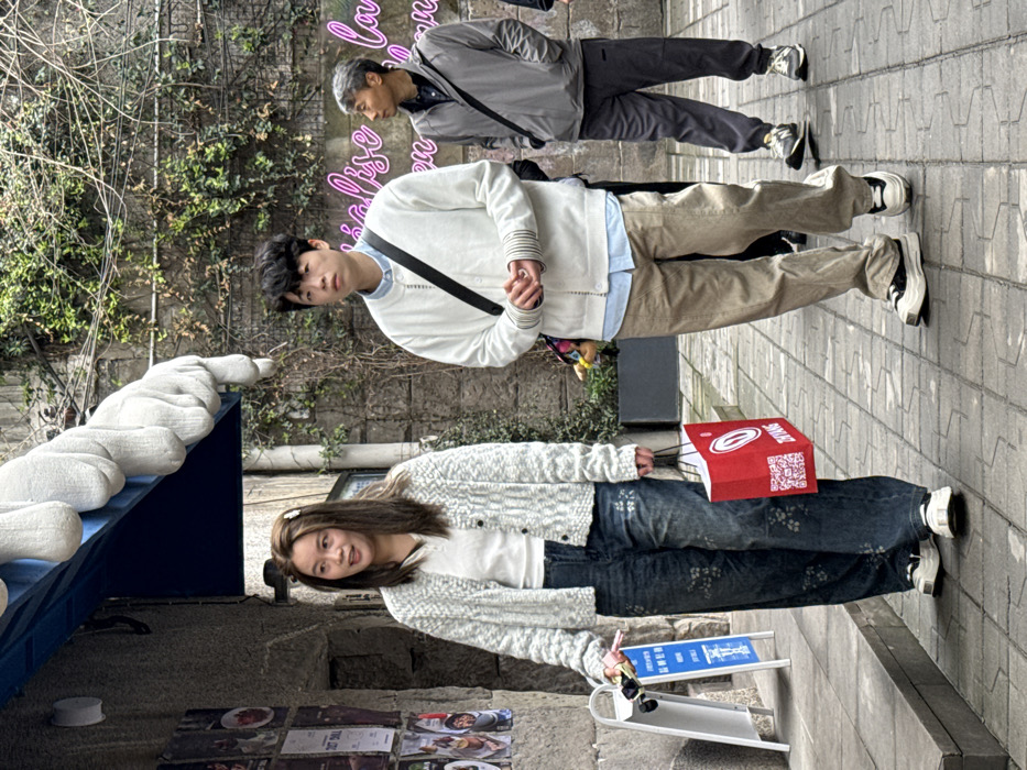 The image depicts an outdoor urban pedestrian area in Yuzhong District, China. Three individuals are walking from left to right on a patterned grey brick pavement. In the foreground left, a woman with long brown hair, wearing sunglasses on her head, a white V-neck top, a light grey textured cardigan, and wide-leg dark denim jeans with a subtle floral pattern, holds a pink object and a black item in her left hand, and a red shopping bag with white text and a QR code in her right hand. She wears white platform sneakers. Her mouth is slightly open. To her right, a man with dark, wavy hair wears a light blue collared shirt beneath an off-white button-up cardigan with striped cuffs, light brown trousers, and black and white platform sneakers. A black cross-body bag is visible over his right shoulder, and his hands are clasped in front. Further back on the right, an older man with short grey hair, wearing a grey zip-up jacket, dark grey track pants, and grey and white New Balance sneakers, also carries a black cross-body bag. To the left, a stone building wall is covered with various posters and a white A-frame sign with blue text. Above the building, a blue awning extends, featuring large white sculptural elements resembling hands. The background includes a stone wall partially covered with green climbing vines and bare branches, with a pink neon sign displaying partial text. A dark planter box with greenery is situated behind the foreground figures.