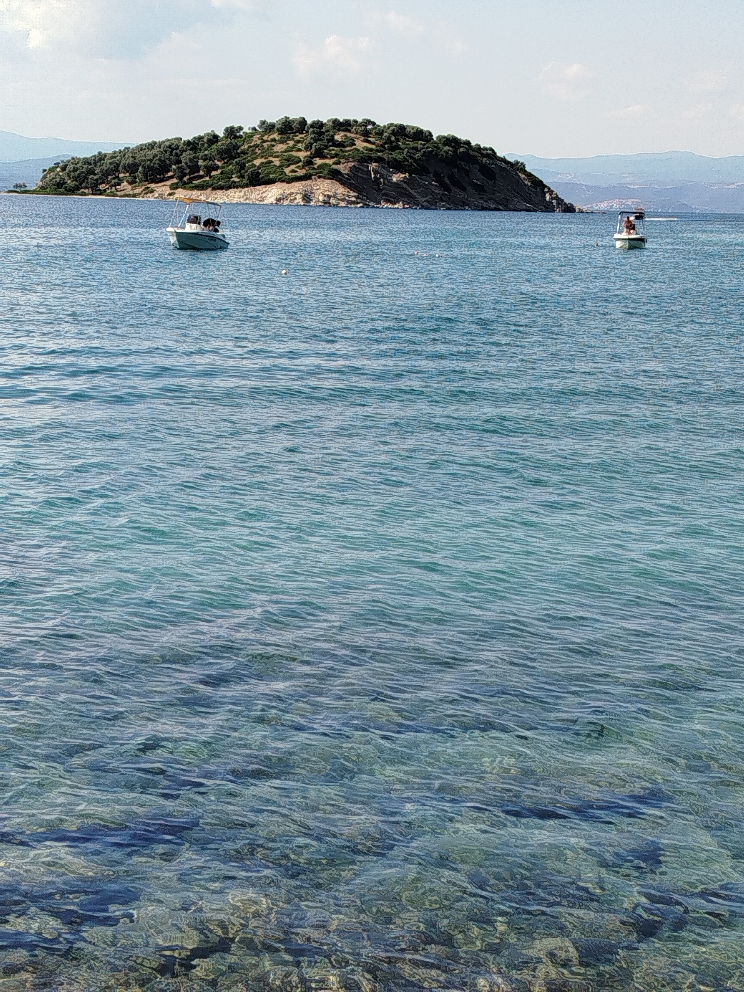 The image depicts a coastal scene in the Sithonia Municipal Unit, Greece. The foreground is dominated by clear blue-green water with visible pebbles on the seabed. In the middle ground, two small motorboats are present on the water. The boat on the left has a light-colored hull and a canopy, with two people visible. The boat on the right also has a canopy, and one person is seated in it. In the background, a tree-covered island rises from the sea, with a rocky slope visible on its side. The horizon is marked by hazy hills under a pale sky with scattered clouds. The overall impression is one of a tranquil summer day at the coast.