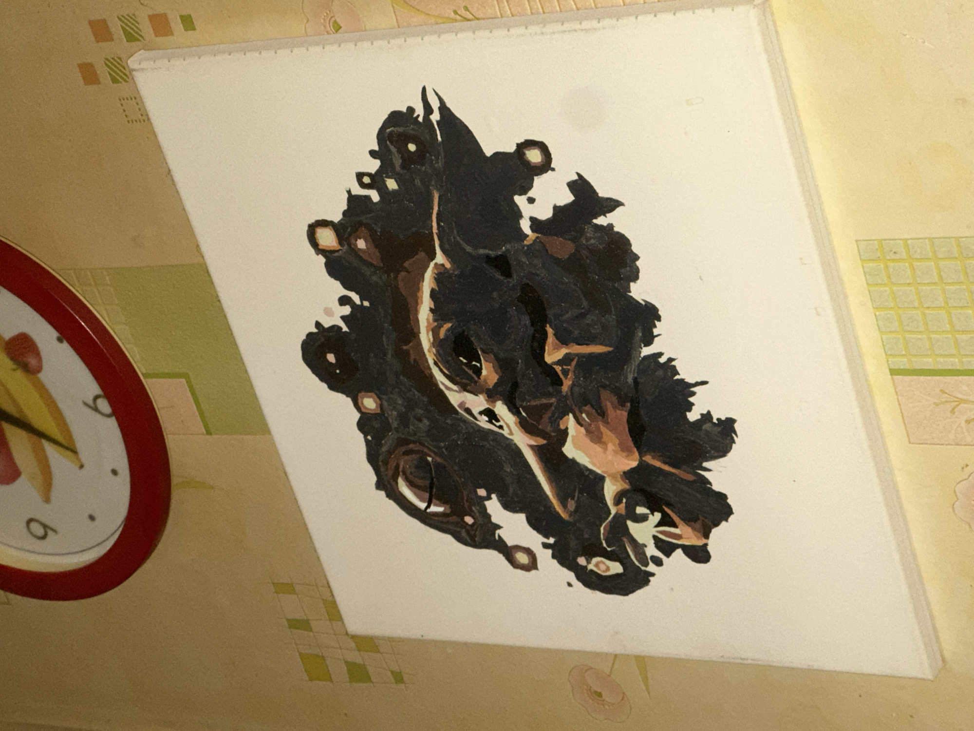 An indoor scene showcases a rectangular canvas featuring a partially abstract painting. The artwork, central to the image, primarily uses black, brown, and off-white tones to depict what appears to be a stylized head of an animal, possibly a dog or a feline, with prominent eye-like shapes and a distinctive snout area.
The canvas is angled, resting against a wall covered in patterned wallpaper. The wallpaper has a light yellowish-beige base. In the upper left corner, there are patterns of small green and orange squares arranged diagonally. Towards the bottom left and center, another section of the wallpaper displays a green and light beige checkered design. Faint, lighter floral patterns are also visible on the wallpaper in the upper right and lower right areas.
In the bottom-left corner of the image, a portion of a circular wall clock with a red rim is visible. The clock face is white, and the numbers '6' and '9' are partially discernible, along with yellow hands, suggesting it's likely around 7:30 or 8:30, though precise time is not clear due to the cropping. The scene is static, providing a glimpse into a domestic interior, possibly in Moscow, Russia, as per the location context. The lighting appears artificial, illuminating the textures of the wallpaper and the painting.