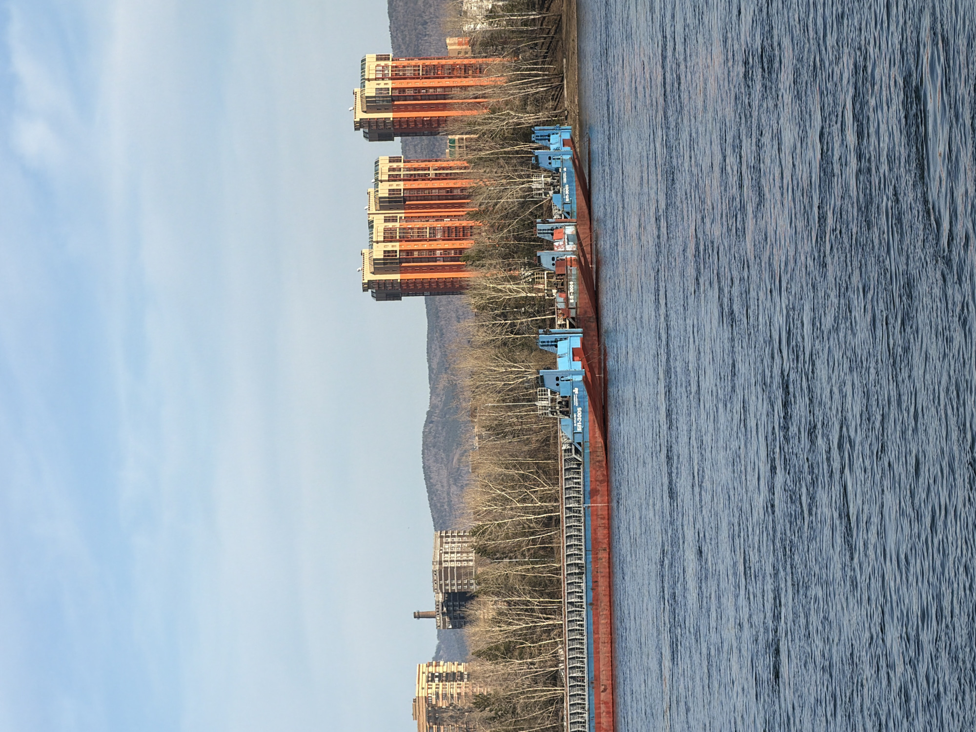 A landscape orientation photograph captures a river scene in Krasnoyarsk, Russia, featuring a barge alongside buildings on the shore. The water of the river covers the right portion of the image. The barge, running parallel to the riverbank, has a rusty red hull and blue machinery mounted on it. Buildings of varying heights and architectural styles line the riverbank. The buildings closest to the barge are residential and commercial. Trees are sparsely interspersed along the shoreline between the buildings and the barge. The sky is light blue and partly cloudy.