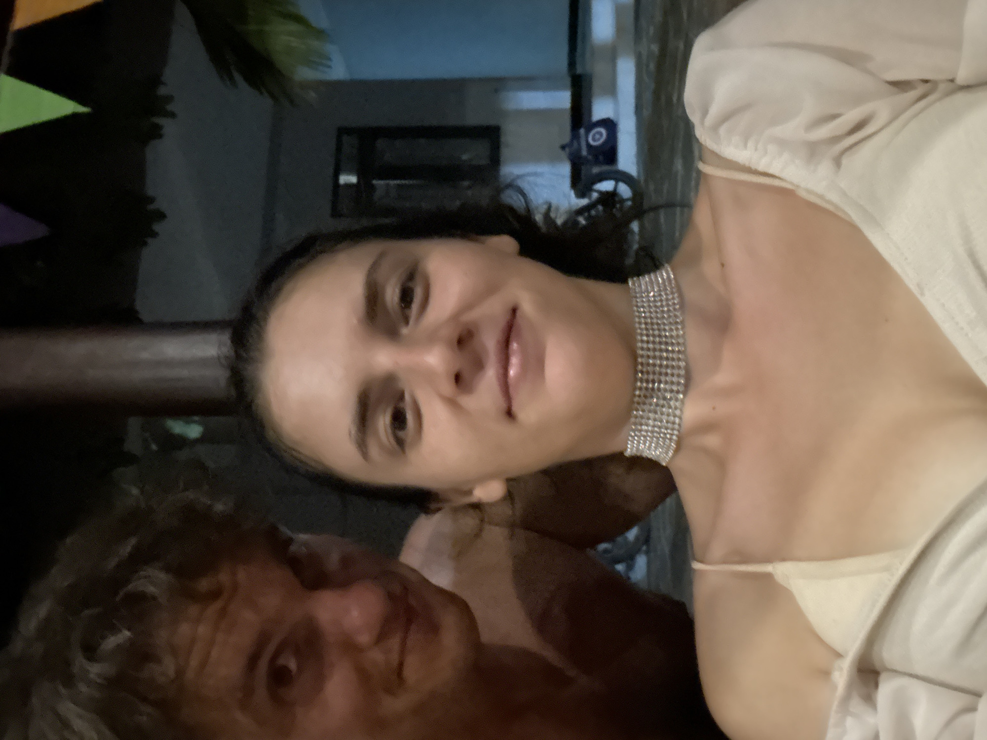 The image depicts two people in close proximity, appearing to be in an outdoor location in Mũi Né, Vietnam. A woman with dark hair and a silver choker is positioned in the foreground, partially in focus. She wears a light-colored top. A man with curly hair is situated to her left, also partially in focus. The background includes a glimpse of building elements and what seems to be a parked vehicle. There is also some hanging decorations of pennants to the upper left of the image.