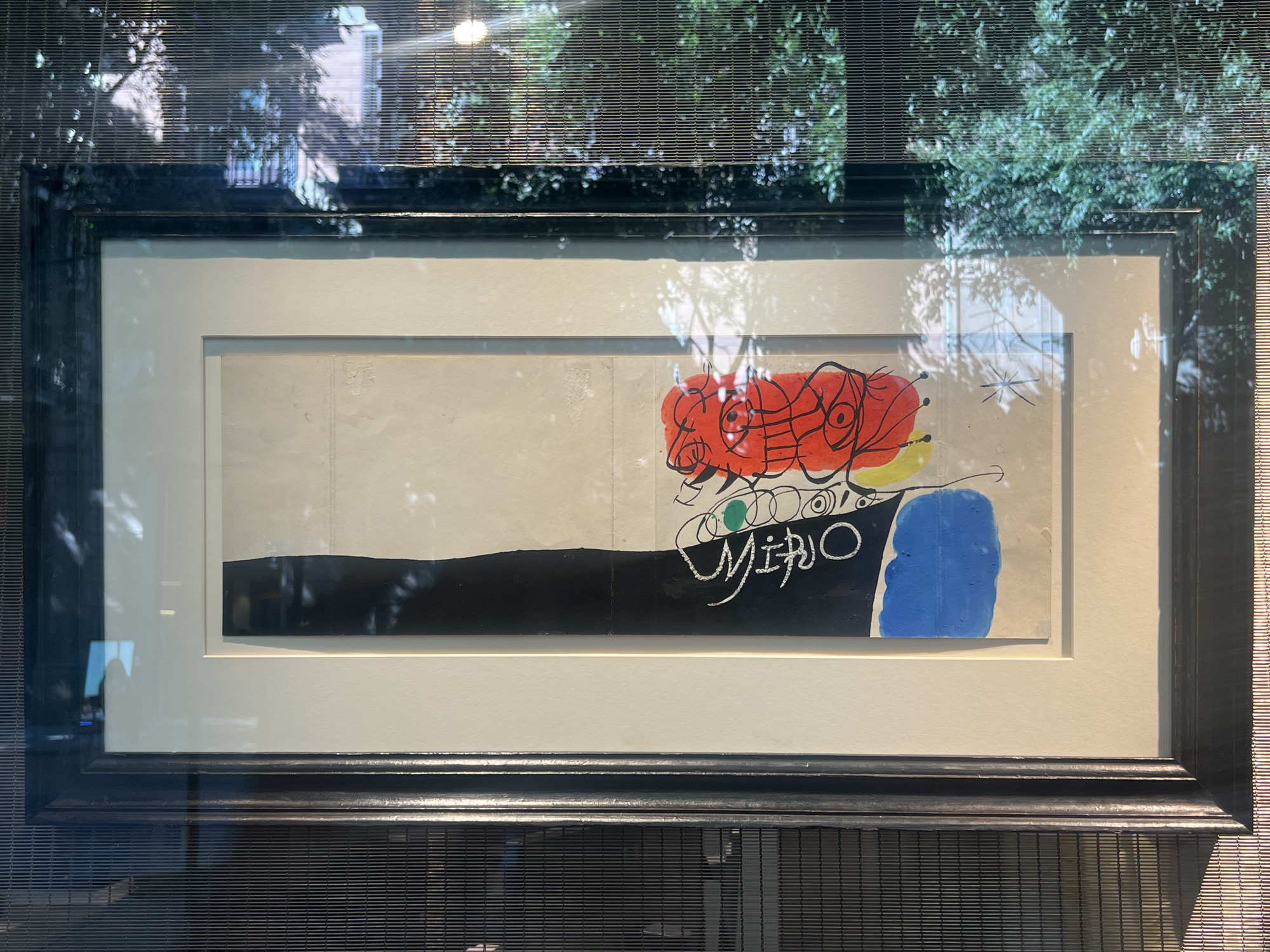 This image displays a framed artwork by Joan Miró, likely a print or lithograph, presented behind glass. The artwork features a predominantly light-colored background with abstract figures and shapes in bold colors, including red, yellow, blue, and black. A prominent red, multi-eyed creature or form dominates the upper central portion of the artwork. Below it, a black horizontal band stretches across the lower part of the composition. The artist's signature, "MIRO", is visible in white script on the black band. The framed artwork is positioned behind a window with a slatted blind, which casts shadows and reflections on the glass, partially obscuring the view of the artwork and its surroundings. Outside the window, trees and buildings are visible, suggesting an urban environment. The overall setting appears to be an interior space, possibly a gallery or private residence, looking out onto an outdoor scene. The style of the artwork is characteristic of Miró's surrealist and abstract expressionist work.
Based on the provided information, this artwork is displayed at the Fundació Joan Miró, located on the mountain of Montjuïc in Barcelona, Spain. The museum houses an extensive collection of Miró's works, and this particular piece, with its distinctive style and signature, is consistent with the art typically exhibited there. The presence of the slatted blind and the reflections in the glass suggest the artwork is either part of a temporary exhibition or a permanent display viewed from a specific angle, possibly within a context that requires light control or has decorative elements that interact with the window. The outdoor scene visible through the window, with trees and hints of urban architecture, aligns with the setting of Montjuïc, a green and somewhat elevated area of Barcelona with significant parkland and views of the city.