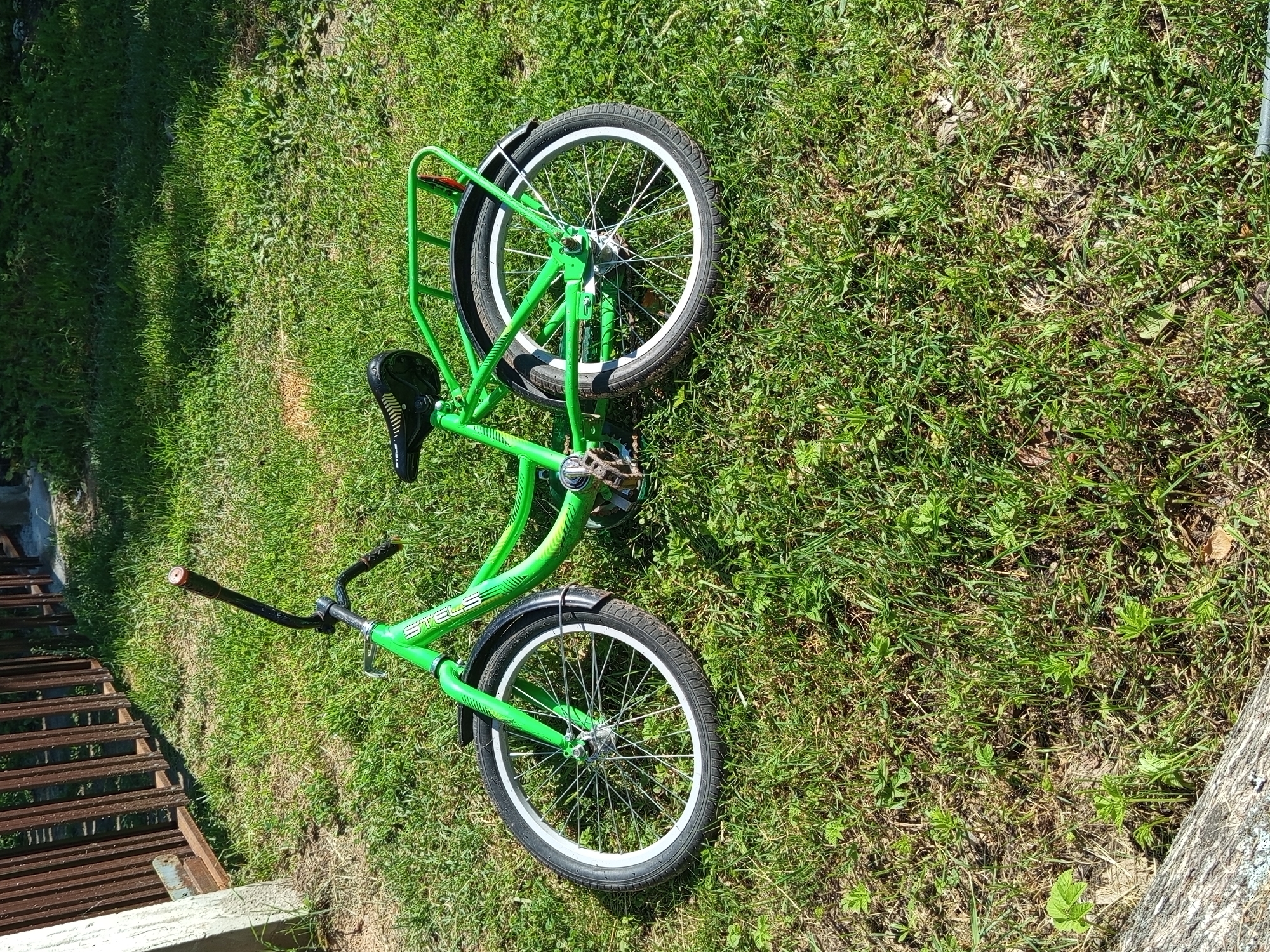 A bright green bicycle is lying on its side on a grassy lawn in Ostrovtsy, Russia. The bicycle is a Stels brand, with a basket on the back and white-rimmed wheels. The handlebars are black and the seat is black with a patterned texture. The bicycle appears to be in good condition. The grass is green and well-maintained, with some patches of dry earth visible. In the background, there is a wooden fence and a concrete structure. The sun is shining, casting shadows on the grass.