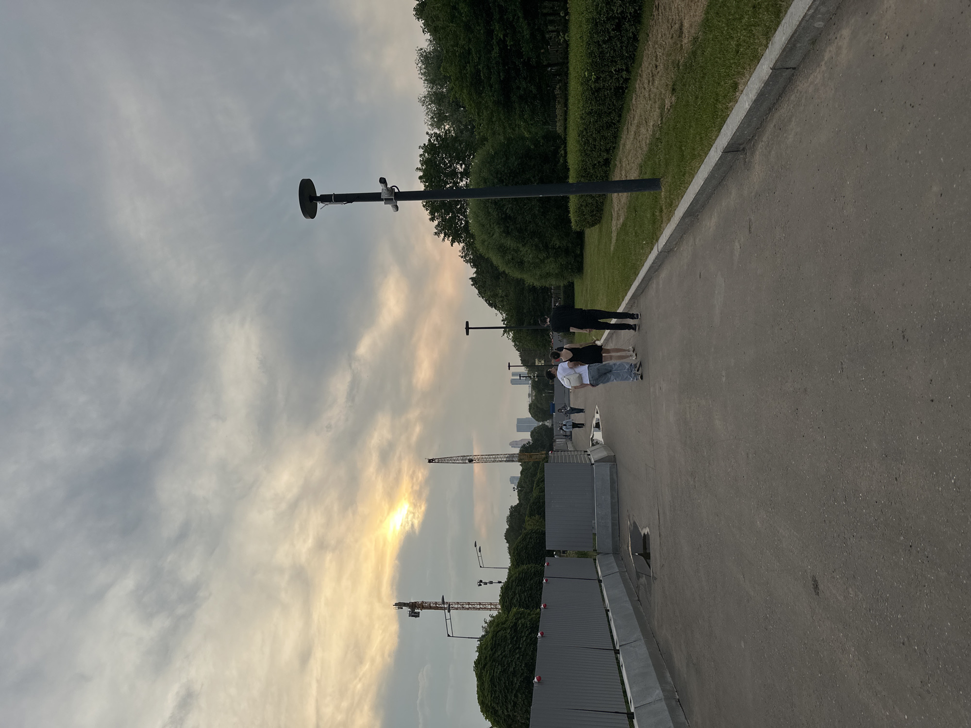 An outdoor, eye-level shot captures a paved pathway on a cloudy day. In the foreground, a man and woman walk away from the camera, followed by a couple, also walking away. A streetlamp with two security cameras stands to the right of the pathway. To the left, a grey metal fence with red accents runs along the path. Beyond the fence, construction cranes and trees are visible. In the distance, skyscrapers can be seen beneath the hazy sky. The ground is paved with asphalt, and patches of grass and bushes line the right side of the path.