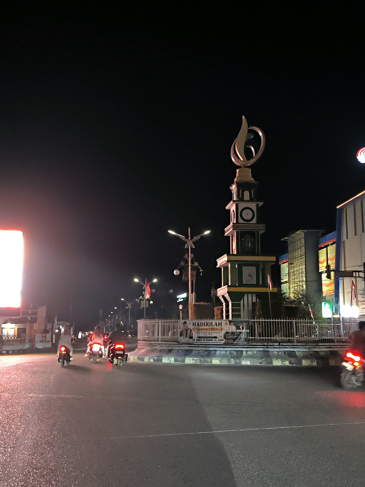 A nighttime street scene in Lhokseumawe, Indonesia, features a prominent clock tower monument with a flame-like sculpture at its peak. The tower, painted in green and white with yellow accents, stands in the center of a roundabout. Several streetlights illuminate the area, and an Indonesian flag is visible on a flagpole near the tower.
To the left of the tower, a large, brightly lit rectangular billboard displays an orange glow. Across the street from the tower, buildings with blue and white facades are visible, some with illuminated signs.
The road in the foreground is wet and reflective, indicating recent rain or moisture. Three motorcycles with riders are visible on the road, moving away from the camera. The motorcycle on the right has a red taillight that is blurred due to motion. The overall atmosphere is of an active urban environment at night, with traffic and illuminated landmarks. A banner with text in Indonesian, likely advertising an event, is partially visible near the base of the roundabout.