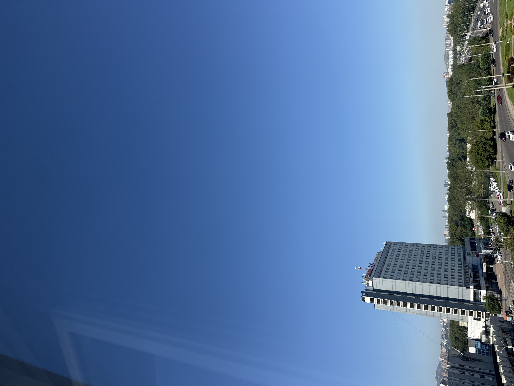 This is a daytime, eye-level, wide-angle photograph taken from a high vantage point overlooking a cityscape. The sky is clear and a vibrant blue, occupying the majority of the frame. In the foreground, to the left, is a portion of a window with a reflection of the sky, creating a subtle distortion.
Dominating the mid-ground is a tall, multi-story building with a light grey facade. This building has a regular pattern of numerous windows. The building appears to be a hotel, as suggested by the signage at the top, which displays the word "MAKS" in red letters, along with Cyrillic text that translates to "hotel" or "hotel complex." The building is slightly tilted in the frame, which could be due to the angle of the photograph or the building's construction.
To the right of the hotel, a wide, multi-lane road is visible with several cars and trucks driving on it. The road curves and appears to be an elevated highway or overpass, with streetlights lining its sides.
The background reveals a dense band of green trees, separating the main buildings from the distant city skyline. The distant city features various buildings of different heights, some appearing residential and others more industrial or commercial. Hills are visible in the far distance.
The overall impression is a view of an urban environment on a clear day, with a focus on a prominent hotel building and the surrounding infrastructure. The specific location is the city of Krasnoyarsk, Russia, as the hotel is the "Maks" Hotel (Hotel Maks), a well-known landmark in the city.