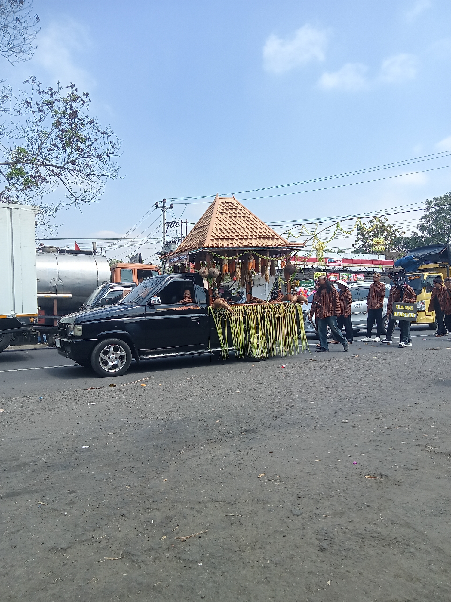 In Urutsewu, Indonesia, on a bright, sunny day with a clear blue sky and scattered clouds, a procession moves along a paved road. A black pickup truck, adorned with a decorative roof made of woven palm leaves and decorated with coconuts and corn, is the central focus. A person is visible driving the truck. Following the truck, a group of men, dressed in matching brown and beige striped shirts and dark pants, walk alongside. One man in the foreground holds a yellow sign that reads "UANG RAKYAT." The procession appears to be taking place in a town, with buildings and power lines visible in the background. Other vehicles, including a white truck with a silver tanker and a yellow truck, are also present on the road. The road surface is asphalt and shows some debris.