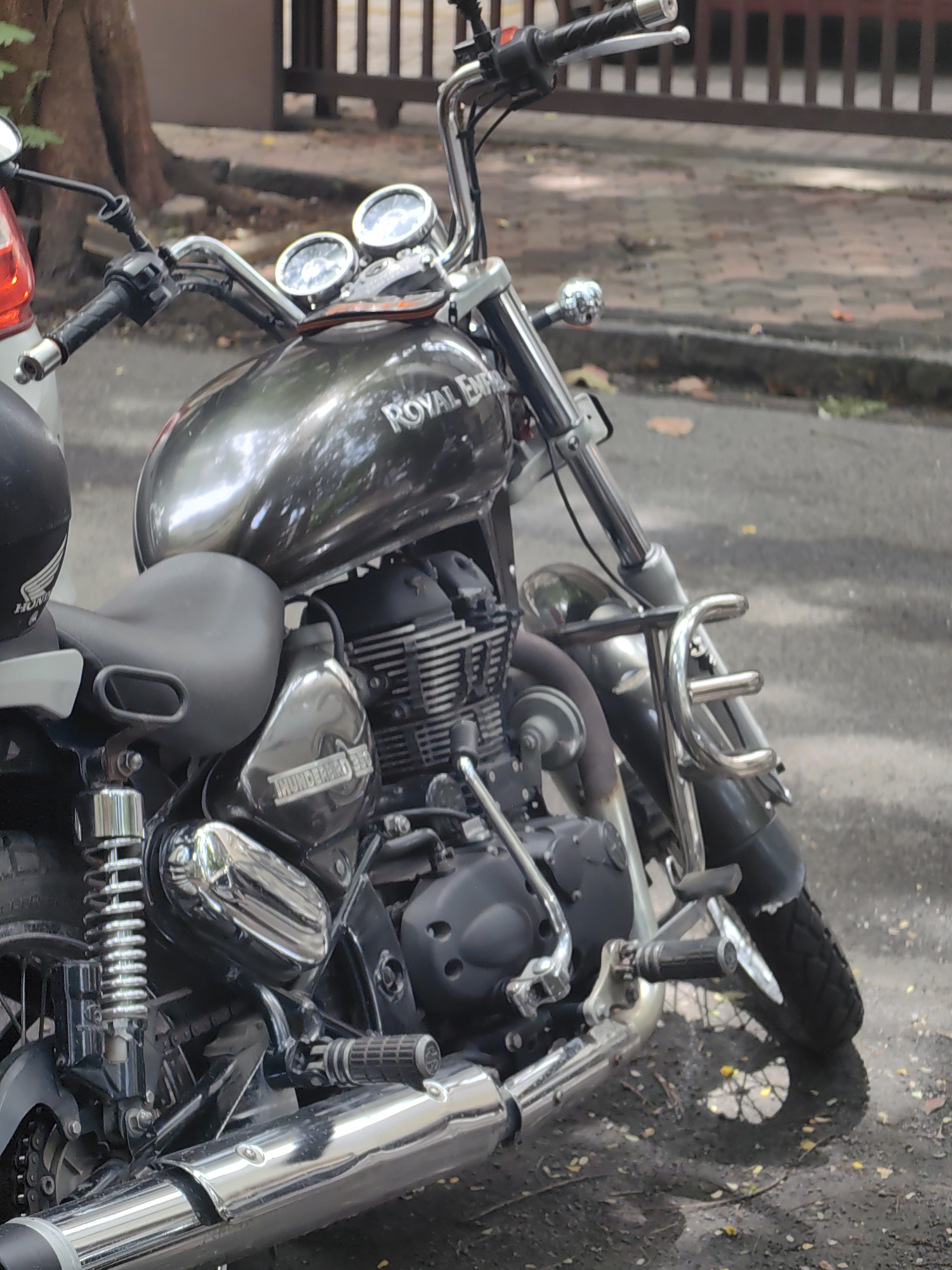An eye-level, medium shot captures a parked Royal Enfield Thunderbird 350 motorcycle on a paved surface, likely a street in Mumbai, India, as indicated by the urban environment and vehicle model common in the region. The motorcycle is predominantly dark grey with chrome accents, featuring a rounded fuel tank with the "Royal Enfield" logo visible, a black leather seat, and a detailed engine block. Chrome handlebars, mirrors, and exhaust pipes gleam, reflecting the ambient light. The front of the motorcycle is equipped with a chrome crash guard. In the background, a blurred tree with a rough bark texture stands to the left, while a wooden fence or gate is visible to the right. The ground beneath the motorcycle is a mix of paved stones and dirt, with fallen leaves scattered around, suggesting a natural or semi-natural urban setting. The lighting appears to be natural daylight, casting subtle shadows and highlighting the motorcycle's metallic surfaces. No people are present in the frame.
