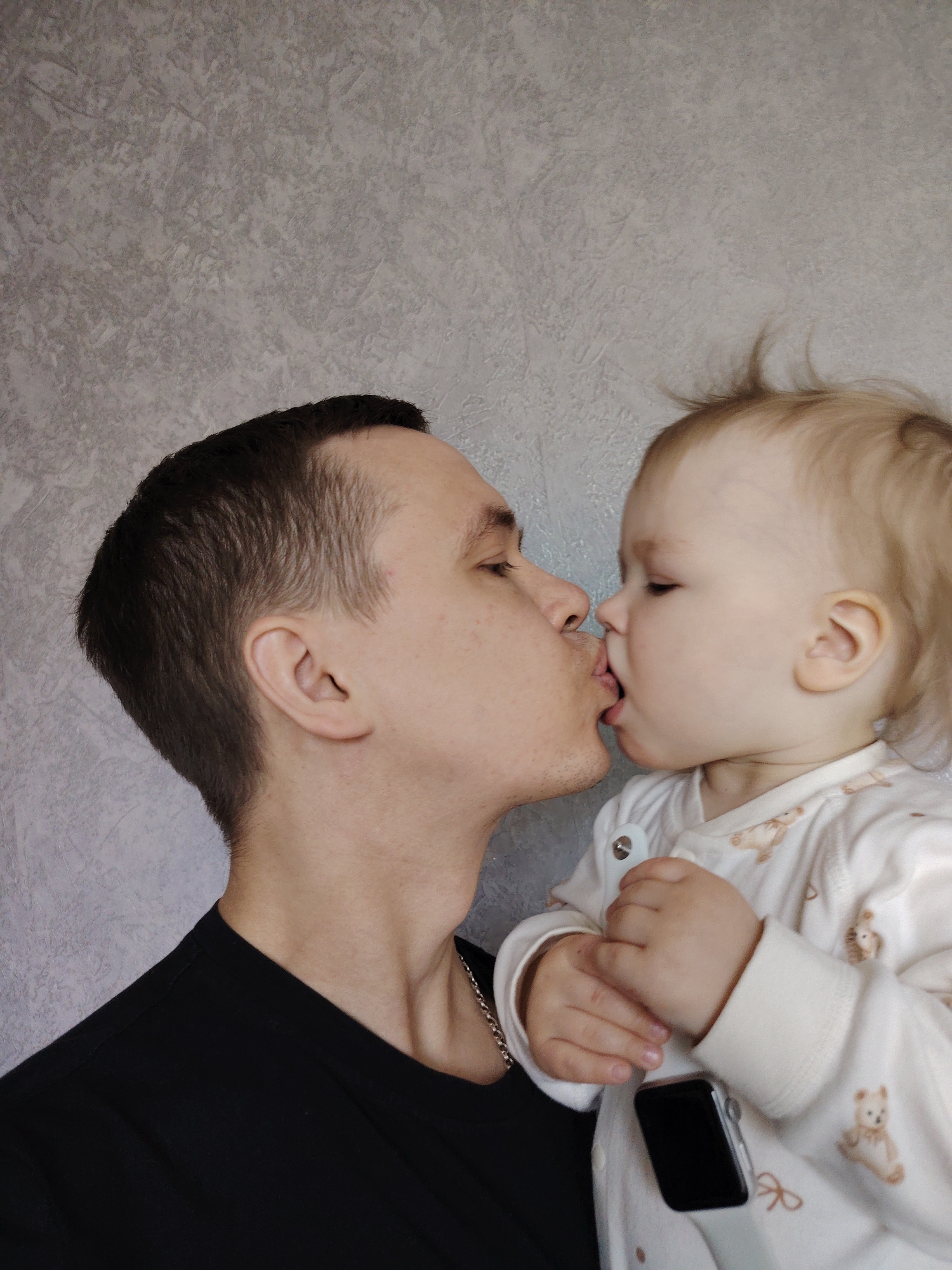 A man and a baby are kissing in front of a gray wall. The man has dark hair and is wearing a black t-shirt with a silver chain around his neck. The baby has blonde hair and is wearing a white onesie with teddy bear patterns. The baby is holding a black watch with a white band. The photo was taken in the city of Chaikovsky, Russia.