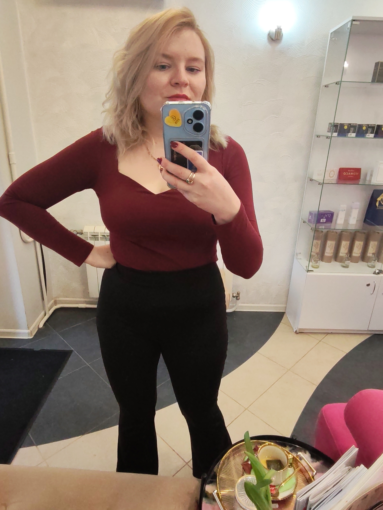 A fair-skinned woman with blonde, wavy hair is taking a selfie in what appears to be a commercial space. She is wearing a maroon, long-sleeved top and black pants. She is holding a blue smartphone with a yellow sticker, likely an iPhone. She has a ring on her finger. The background reveals a tiled floor with a black and white pattern, a white retail display case with various products, and a pink chair. A small table with a cup and papers on it is visible in the foreground. Based on the style of interior decoration and available merchandise, it's plausible this photo was taken in a salon or boutique in a city like Izhevsk, Russia, although this is not confirmed.
