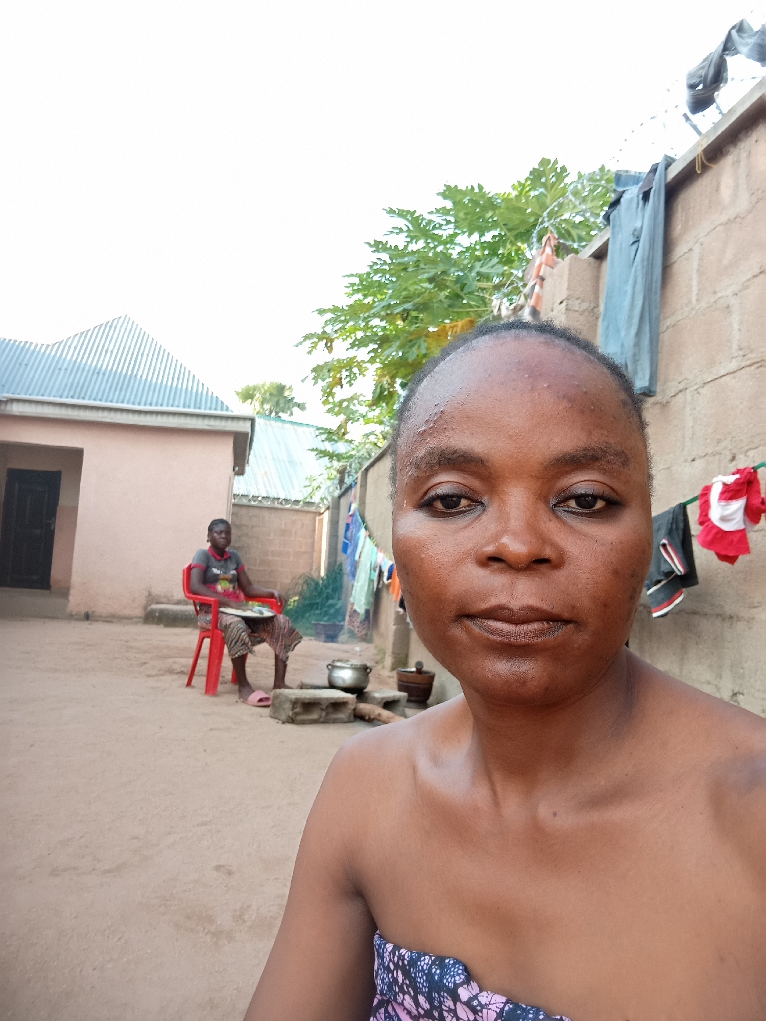 The image shows a close-up of a woman's face in the foreground. She has dark skin and appears to be outdoors. In the background, a person is sitting on a red plastic chair in a dusty courtyard. There are buildings with corrugated metal roofs, a brick wall, and laundry hanging on a clothesline. A pot is visible on the ground near the person in the chair. The overall impression is of a simple, rural or semi-rural setting, possibly in a village or compound in a city like Kason, Nigeria.