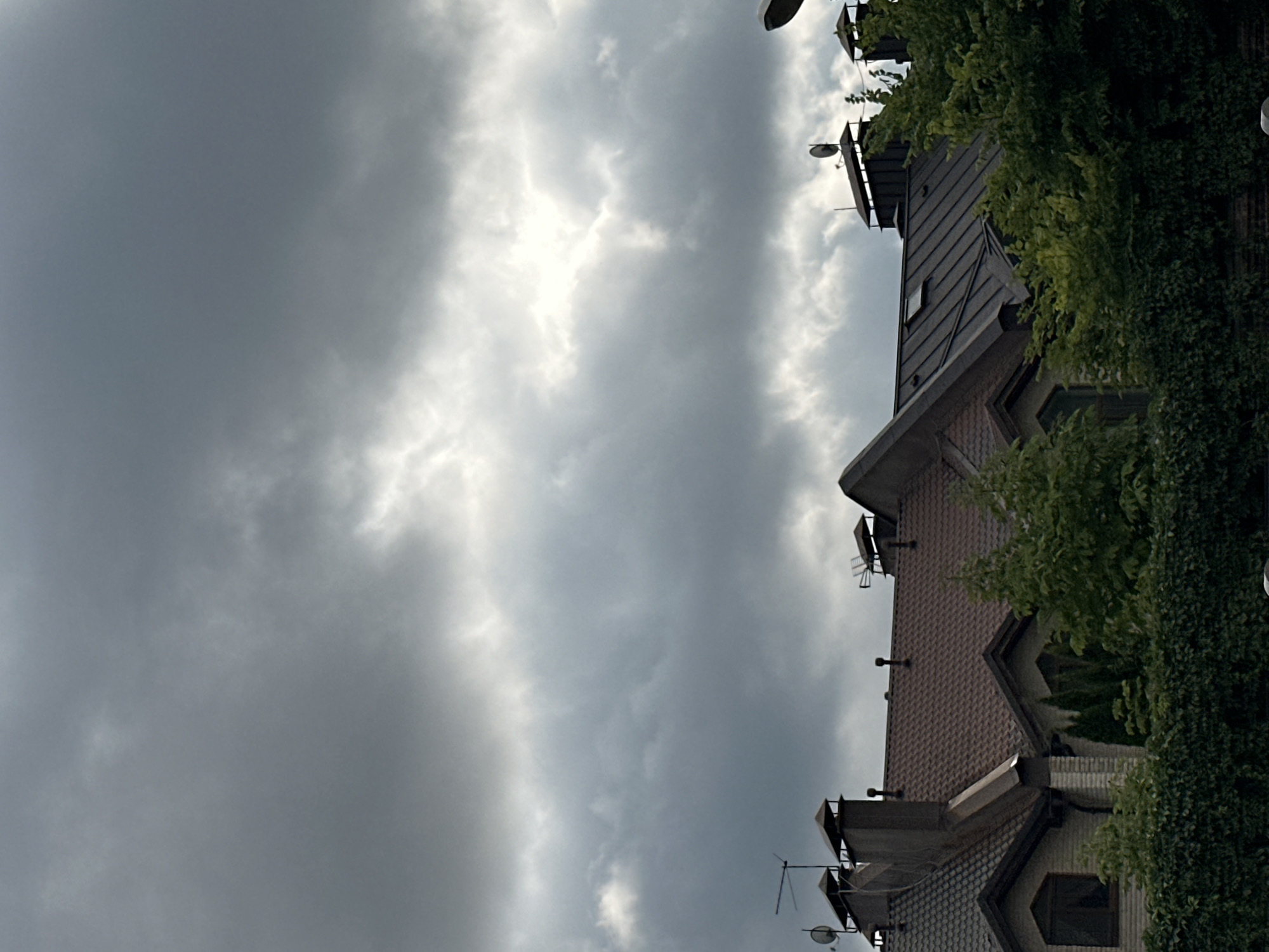 The image shows the rooftops of several houses with dark grey, cloudy skies above. The houses have tiled roofs, some with chimneys, satellite dishes, and antennas. Trees and dense greenery are visible in front of the houses. The scene appears to be a residential area in Yurlovo, Russia.