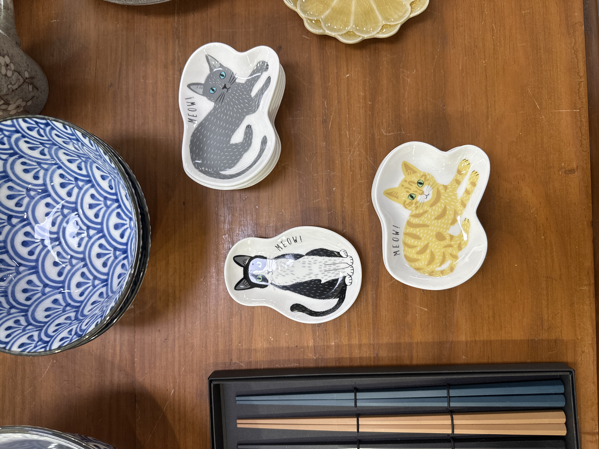The image shows a collection of ceramic dishes and chopsticks laid out on a wooden surface. In the top left is a stack of blue and white patterned ceramic bowls. To the right of the bowls are three small, irregularly shaped white ceramic dishes, each with a different cat illustration and the word "MEOW!". The top dish depicts a grey cat, the middle dish shows a black and white cat, and the bottom dish features a ginger cat. To the left of the cat dishes is a black box containing a set of chopsticks, with two pairs visible: one pair appears to be a light brown wood, and the other pair is a dark teal color. On the far right of the image, a portion of a yellow, textured ceramic dish is visible. The overall arrangement suggests a display of tableware or decorative items. There are no people or actions depicted. The environment appears to be indoors, with the wooden surface suggesting a table or countertop. The image was taken in the city of Bellingen, Australia.