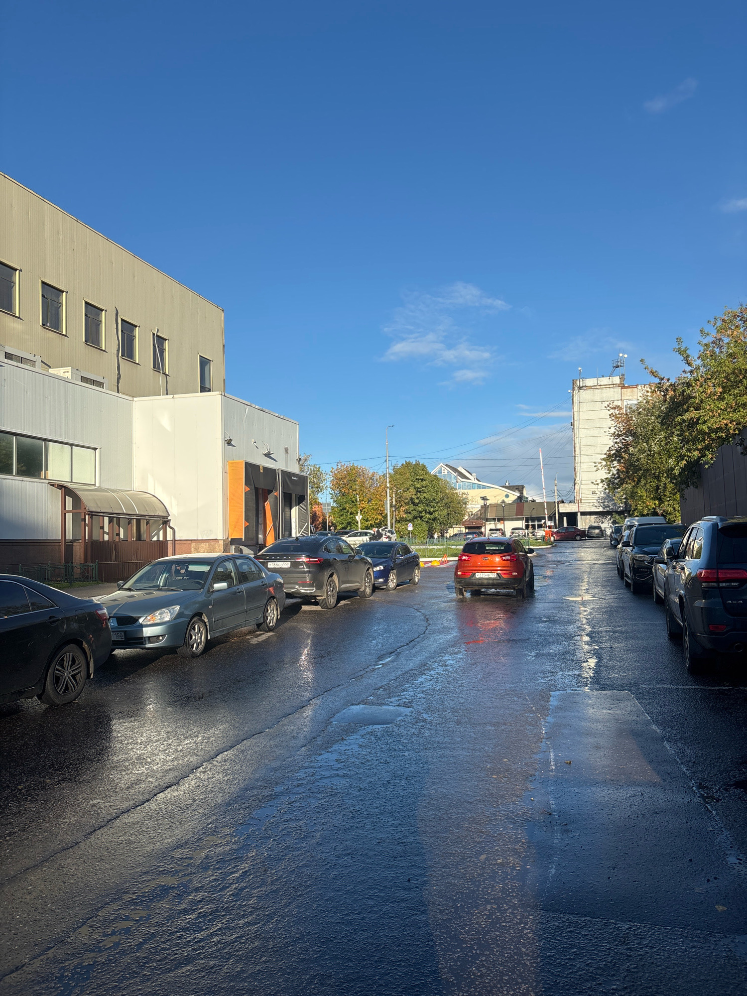 An external, eye-level, medium shot captures a street scene in Sergiyev Posad, Russia, under a bright, clear blue sky with a few wispy clouds. The wet asphalt of the street reflects the sunlight and the surrounding environment. Parked cars line both sides of the road. On the left, a light gray industrial-style building with numerous windows dominates the foreground. Adjacent to it is a white structure with orange accents and a covered entrance. To the right, a taller, weathered concrete building with a white upper section stands, partially obscured by trees with autumn foliage. The foreground on the right shows a line of parked SUVs. The overall impression is that of a commercial or industrial area, likely on a day following rain.