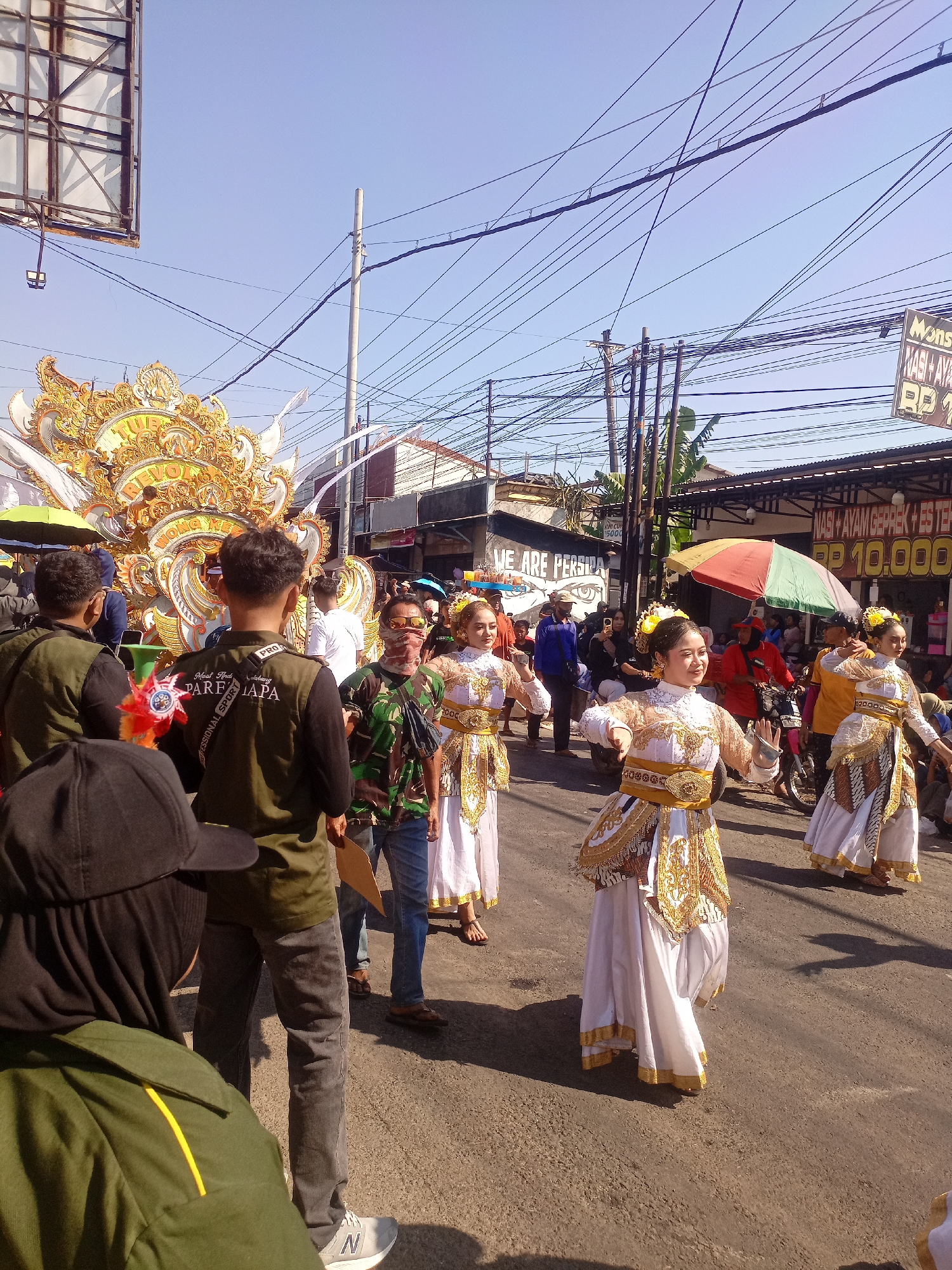This image depicts a parade or festival taking place on a street in Margoyoso, Indonesia. The foreground features several individuals, including those in dark green vests and one wearing a black baseball cap and hijab. In the middle ground, a group of women in traditional white and gold attire are performing a dance. They have elaborate gold headpieces and are smiling. Behind them, a large, ornate float adorned with gold and white decorative elements is visible. The street is lined with buildings and shops, some with signs indicating food sales. A colorful umbrella is open, offering shade. Overhead, a complex network of power lines crisscrosses the sky, and a large billboard structure is also present. The overall atmosphere appears festive and sunny, with spectators lining the street.
