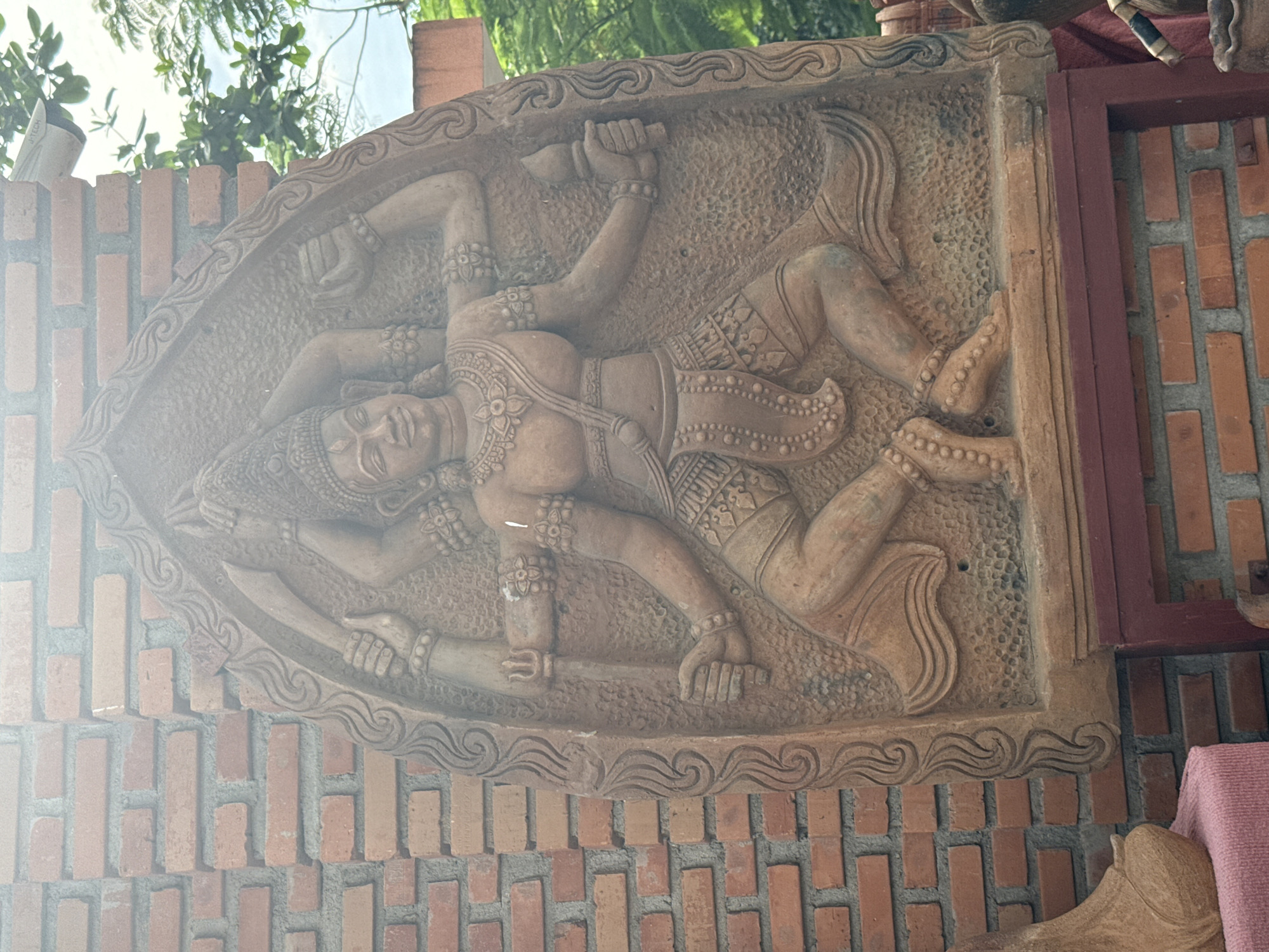 The image shows a bas-relief carving of a multi-armed figure, possibly a deity or mythological being, mounted on a red brick wall. The figure is depicted in a dynamic pose with ornate jewelry, headdress, and clothing details. The carving itself is framed by a decorative border. A red frame is visible to the right of the carving. The wall is part of a larger structure, possibly a building or temple in Bac Nha Trang Ward, Vietnam. Green foliage and sky are visible in the upper left corner.