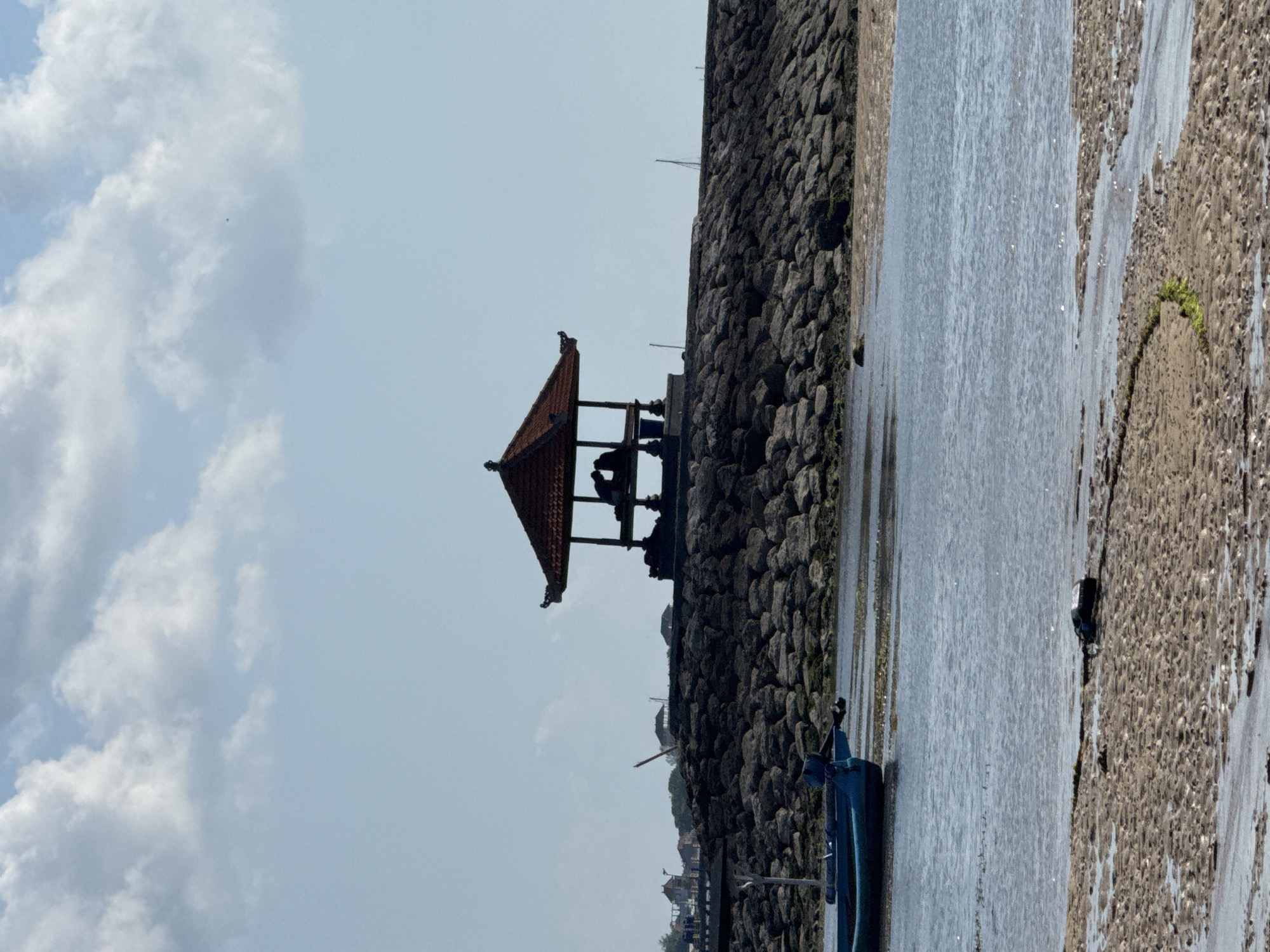 The image depicts a tranquil scene at a beach in Denpasar, Indonesia. In the foreground, a sandy shore with tidal pools and seaweed is visible, with a dark object, possibly a piece of debris, resting on the sand. A calm body of water separates the shore from a rocky breakwater. Two blue boats are moored in the water near the breakwater.
Dominating the midground is the breakwater, constructed of large, dark rocks, which leads to a traditional Balinese pavilion or gazebo. The gazebo features a multi-tiered, tiled roof in a rich reddish-brown color, supported by dark pillars. Silhouetted within the gazebo are two individuals, seemingly seated, their interaction not clearly discernible due to the backlighting.
In the background, the sky is a pale blue, partially obscured by scattered white clouds. To the left of the gazebo, a distant view of buildings and vegetation suggests a coastal settlement. The overall atmosphere is serene, with a focus on the natural elements and the architectural feature of the gazebo.