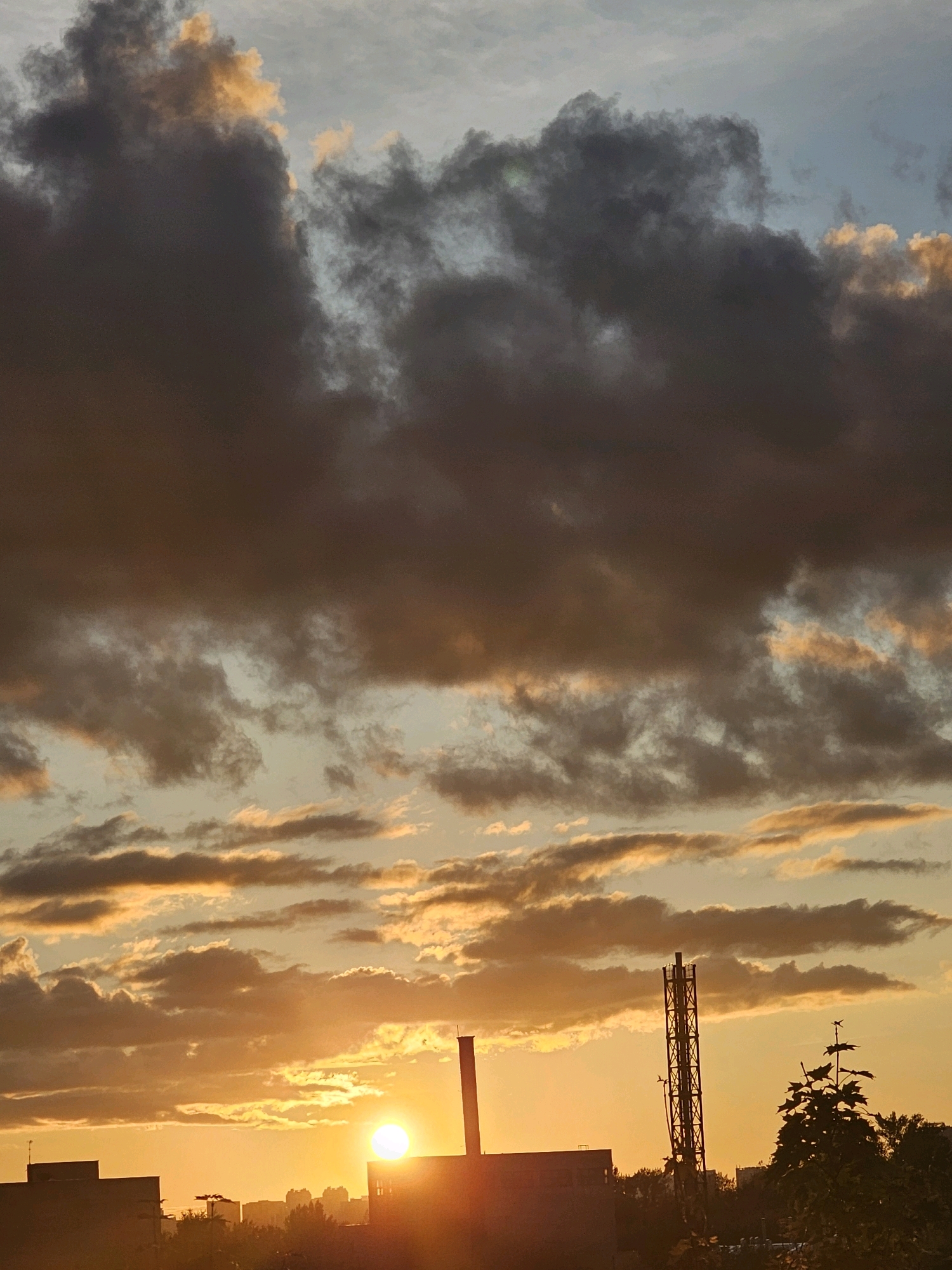 A sunset view over the city of Khimki, Russia, is captured in this image. The sun, a bright orb of light, is low on the horizon, casting a warm orange glow across the scene. Dark, dramatic clouds with streaks of gold and orange fill the sky, creating a striking contrast with the lighter blue sky above. In the foreground, the silhouettes of buildings and industrial structures are visible. A tall, lattice-like tower stands prominently on the right, while a smokestack rises from a building on the left. The silhouettes of trees and vegetation can be seen beneath the structures, adding a touch of nature to the urban landscape. The overall impression is one of a dramatic and picturesque end to the day in a Russian city.