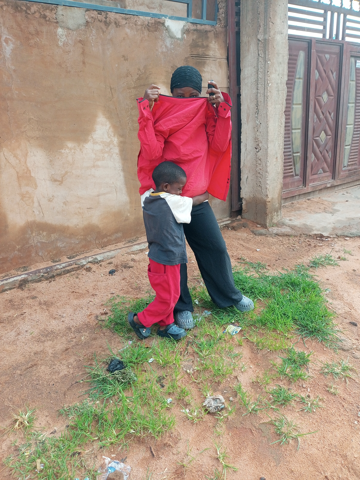 In the city of Tempure, Nigeria, a woman wearing a black headscarf and a red shirt is partially obscuring her face with the shirt as she holds a dark object to her face, possibly a phone. She is standing next to a young child who is facing away from the camera, with their eyes closed, and is wearing a grey and white shirt with red pants and black Crocs. They are standing on a dirt ground with patches of grass. Behind them is a textured, tan-colored wall and a building with large wooden doors. The scene appears to be outdoors, with natural light.