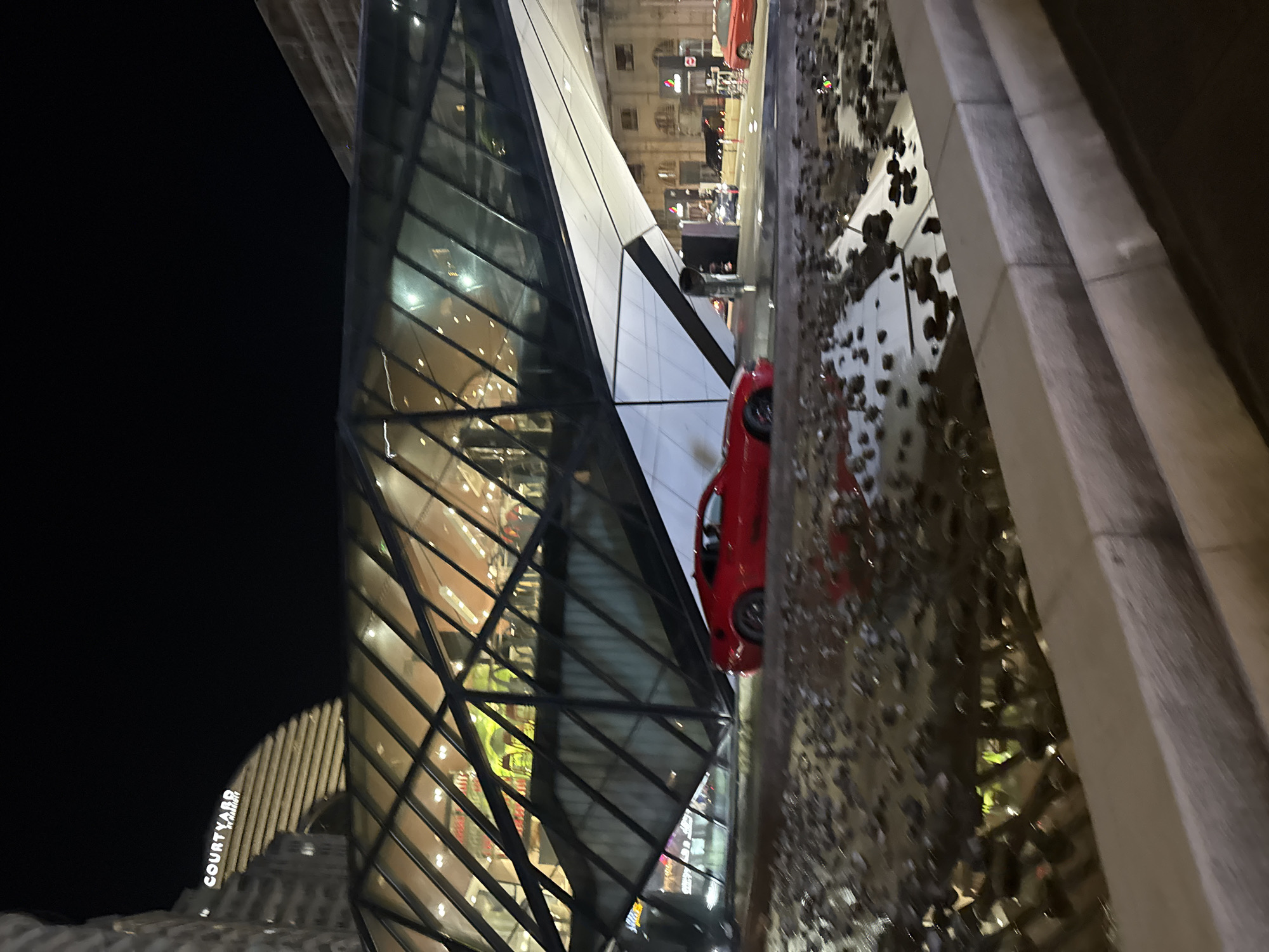 Captured at night, the image presents a sharp upward perspective of modern architecture adjacent to a street scene. A contemporary building with a distinctive angular design dominates the left, featuring a crisscrossing pattern of dark metal beams enclosing reflective glass panels, suggesting multiple stories within. A red automobile is positioned on the street in the middle, partially obstructed by a sculptural water feature adorned with an arrangement of dark, possibly metal, embellishments. The environment seems urban, with a second building marked "Courtyard" visible on the lower left, adding a layer of architectural context. The overall image conveys a sense of urban modernity, juxtaposing architectural design with urban infrastructure.