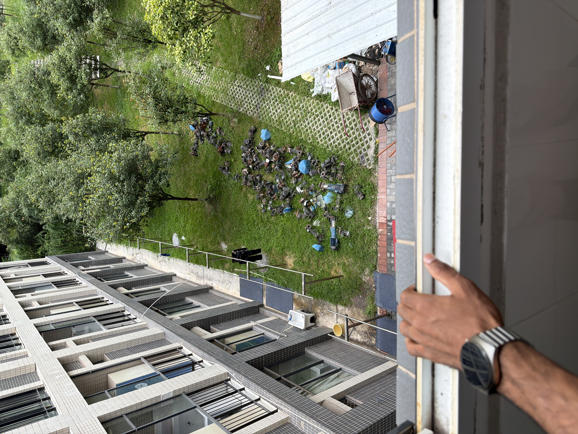 In Jiangjin, China, an outdoor scene unfolds adjacent to a modern, multi-story residential building with a tiled facade and numerous balconies. A person's hand, wearing a silver-banded watch, rests on the windowsill of a lower floor, suggesting the image was taken from within the building. Below the building, a grassy area is interspersed with trees and a permeable paver pathway. A collection of what appear to be discarded pumps and blue plastic objects are scattered across the lawn, near a person wearing dark clothing who is standing beside a white plastic basin and a yellow bucket on a small table. Further along the pathway, a wheelbarrow, a blue industrial fan, and various other items are visible, including sacks of material and electrical equipment near a corrugated metal awning. The overall impression is one of a cluttered outdoor space, possibly a communal area or storage yard, adjacent to residential units.