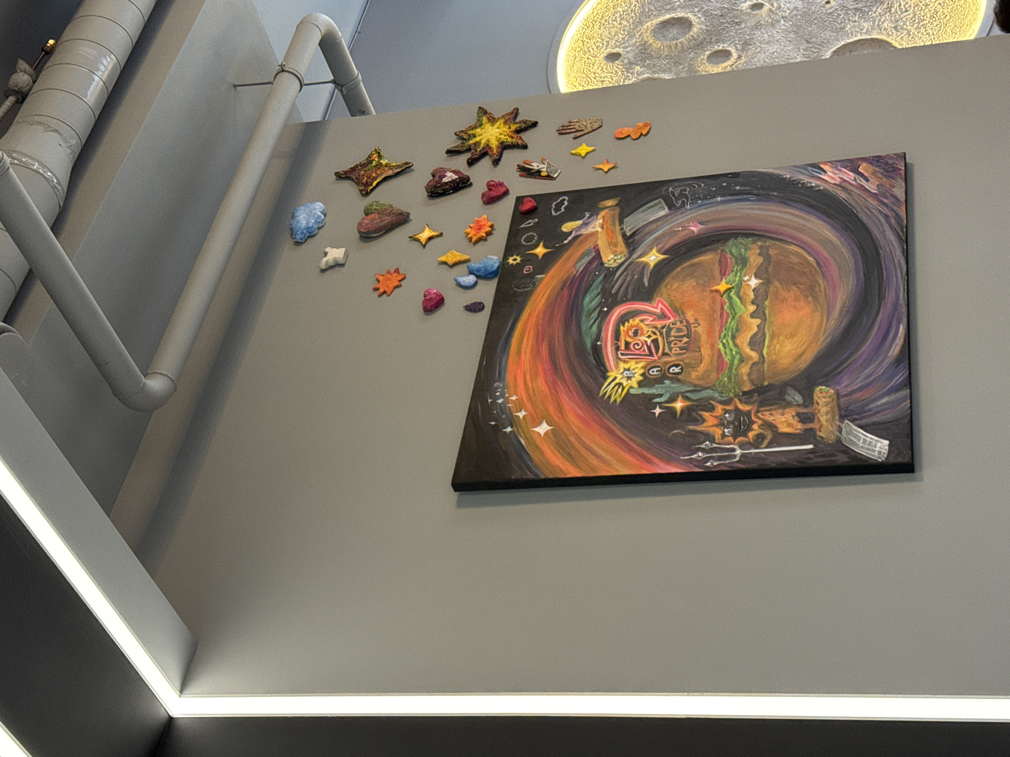The image shows a close-up of a grey wall in what appears to be a restaurant in Saint Petersburg, Russia. On the wall, there is a colorful, swirling painting of a hamburger in a cosmic vortex. Above the painting, a collection of small, colorful, three-dimensional art pieces are attached to the wall, including hearts, stars, and abstract shapes. Overhead, exposed pipes and ductwork are visible, illuminated by strip lighting along the top edge of the wall. To the right of the painting, a portion of a large, textured decorative moon is visible, illuminated by a warm light.