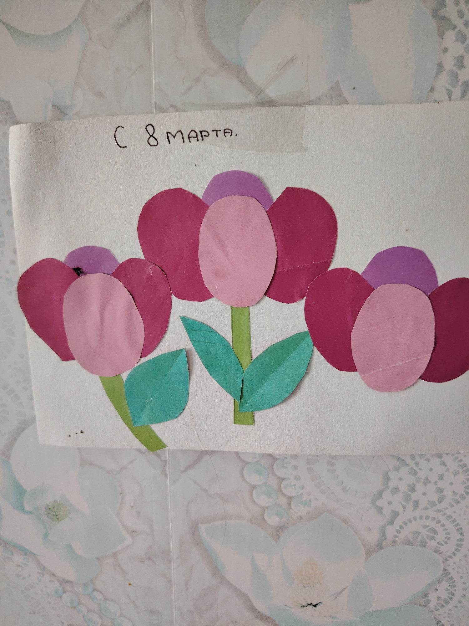 The image displays a handmade paper craft, likely a greeting card, featuring three stylized flowers. The flowers are created from paper cutouts in shades of pink, magenta, and purple, arranged on a white background. Each flower has a central light pink or dark pink petal, surrounded by magenta petals, and topped with a purple petal. Green paper cutouts form the stems and leaves beneath the flowers.
At the top of the white paper, handwritten in black, is the text "С 8 МАРТА.", which translates from Russian as "Happy 8th of March" or "To 8th of March," indicating a celebration of International Women's Day.
The craft is placed on a light-colored, patterned surface, possibly wallpaper or a table covering, adorned with a delicate white and light blue floral and lace-like design. The image captures a static view of the finished craft, conveying a sense of a personal, handmade gift intended for a celebratory occasion. There are minor imperfections on the paper, such as small smudges or creases. This craft was made in Сусады-Эбалак, Russia.