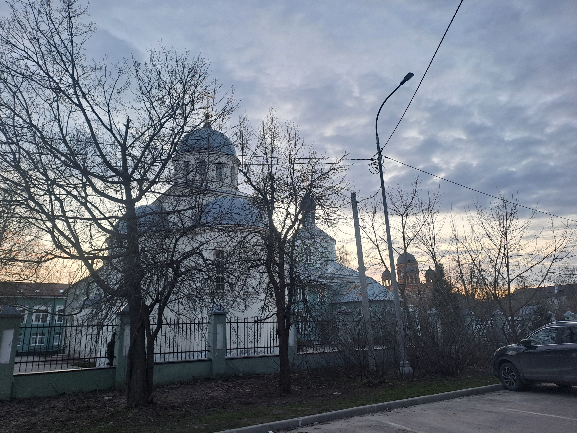 The image depicts a scene in Elektrostal, Russia, featuring multiple buildings, most notably a white church with blue domes and a golden cross atop the largest dome. A second church is visible with a red dome. The buildings are partially obscured by bare trees, typical of a winter or early spring setting. A black car is parked to the right of the frame. A street light pole is situated in the middle ground, with wires extending from it. The sky is overcast with a mix of grey and white clouds. A section of a building with green exterior paint and a black metal fence is visible along the bottom left of the image. The ground is mostly paved with sections of grass, some of which are covered with fallen leaves. The overall composition suggests a quiet, urban environment.
