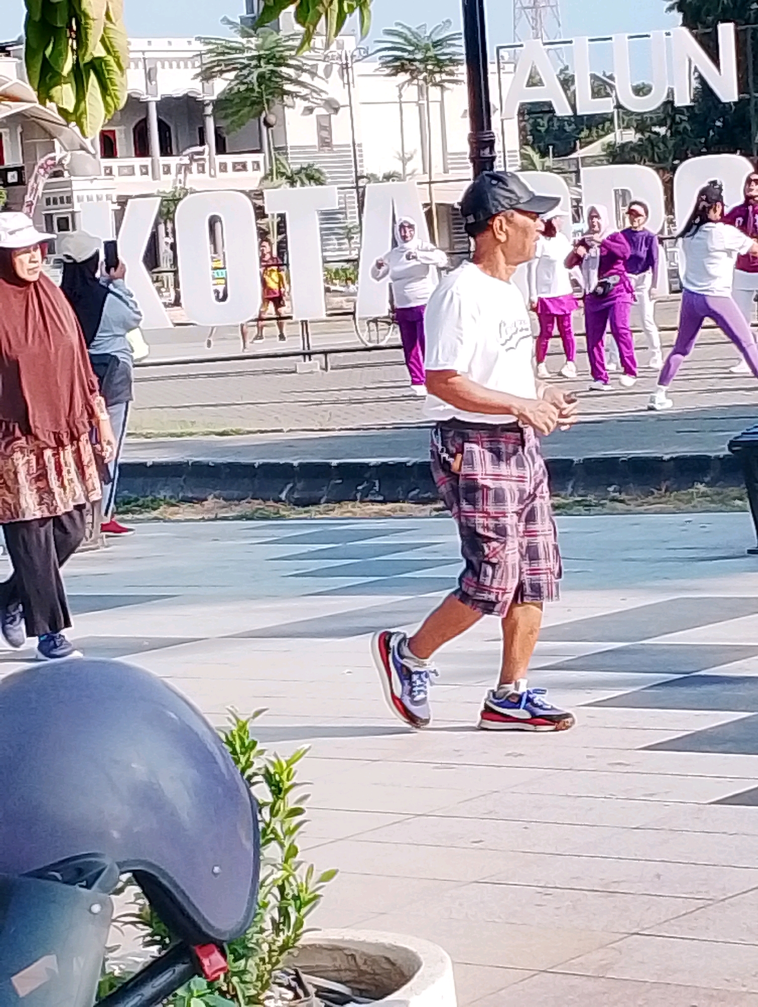 The image depicts an outdoor scene in what appears to be a public square or park in Probolinggo, Indonesia. In the background, large white letters spell out "KOTA" on the left and "ALUN" on the right, suggesting a sign for "Alun-Alun Kota," a common designation for town squares in Indonesia.
In the foreground, a man wearing a black cap, a white t-shirt, and plaid shorts is walking towards the right side of the frame. He has his hands clasped in front of him. To his left, a woman wearing a brown hijab and a patterned tunic is also walking. Behind them, a group of people, primarily women dressed in athletic wear (purple tops and bottoms, or white tops and purple bottoms), appear to be participating in a fitness activity, possibly exercising or stretching. Some of them are holding their hands up in unison. The ground is paved with light-colored tiles, and there are some green plants visible. A portion of a dark motorcycle helmet is in the lower left corner of the image. The overall atmosphere seems to be one of community activity and recreation.