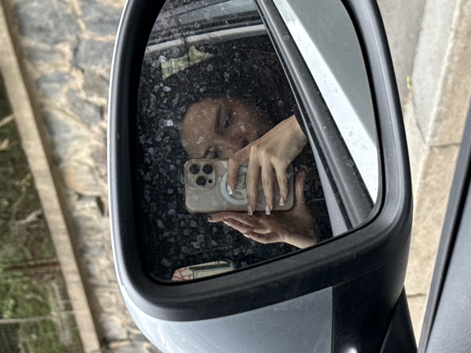 In a reflection within a car's side mirror, a young woman with long, dark hair is captured holding a smartphone to take a selfie. Her face is partially obscured by the phone, and she is wearing dark clothing. The mirror's surface is visibly dirty with numerous smudges and specks. The background of the reflection shows the interior of a car, including a window and what appears to be a building or wall. The exterior visible around the mirror frame is blurred, with hints of a stone wall and greenery, suggesting an outdoor urban or suburban setting, potentially in Prague, Czechia.