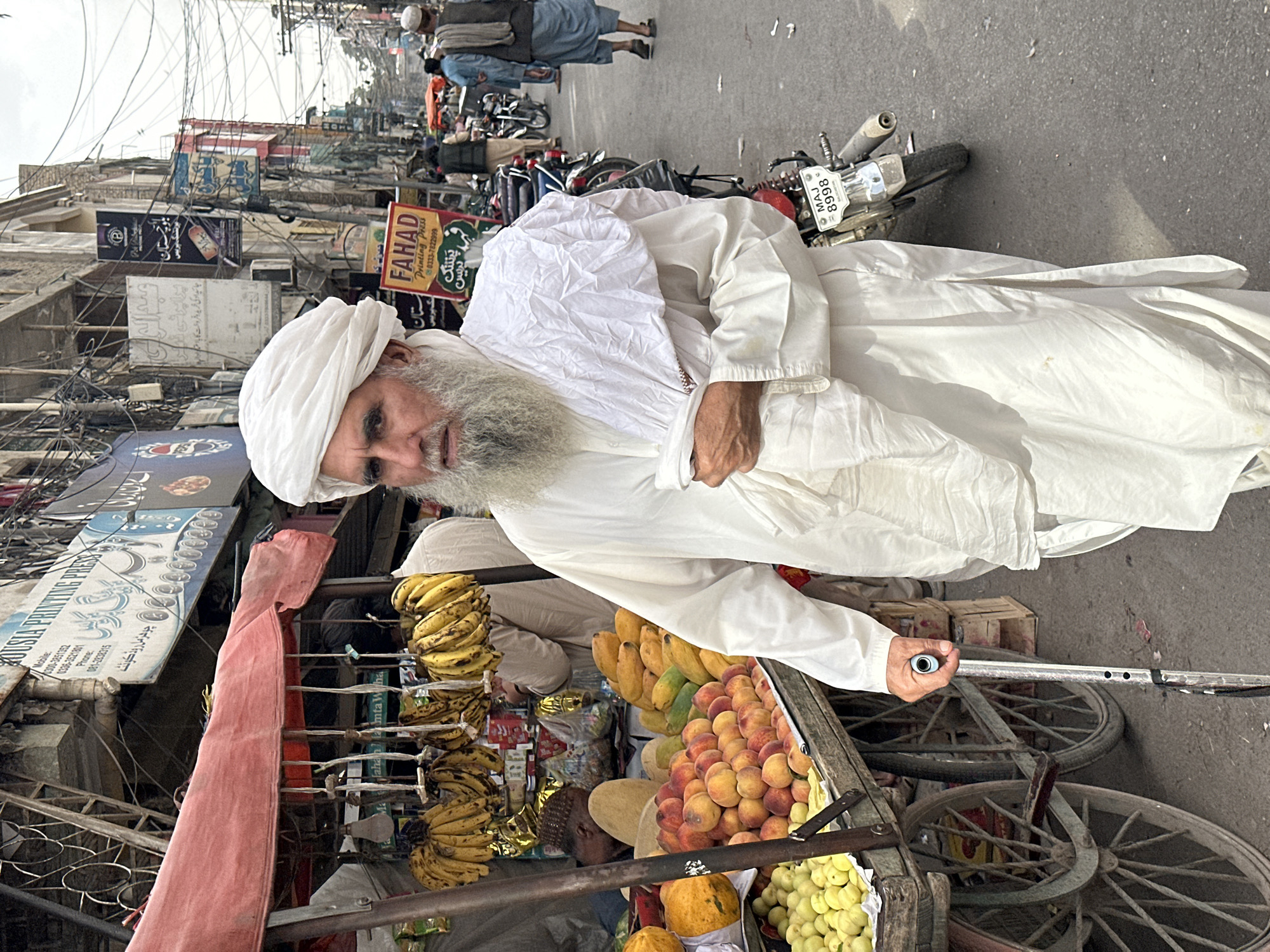A man with a white beard, wearing a white turban and traditional Pakistani clothing, is walking with a cane in the street of Quetta City Tehsil, Pakistan. He is pushing a cart loaded with fruit: bananas, peaches, papayas, and grapes. The street is a bustling market with many vendors, motorcycles, and pedestrians. The buildings lining the street are old and have many signs. The sky is overcast. There are many electrical lines strung across the street.